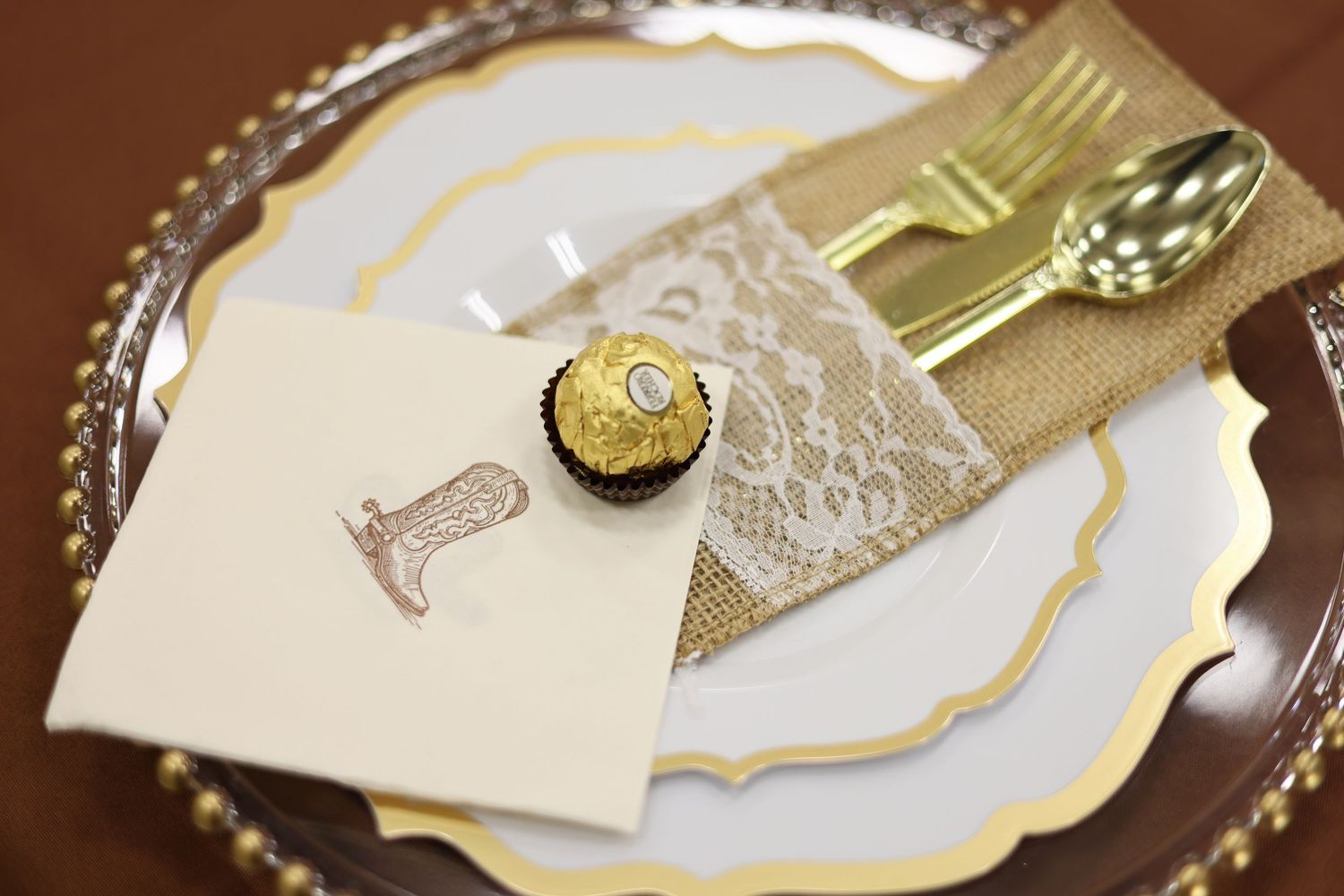 Gold-rimmed plate with burlap lace cutlery holder, Ferrero Rocher chocolate, and elegant place card for a formal event.