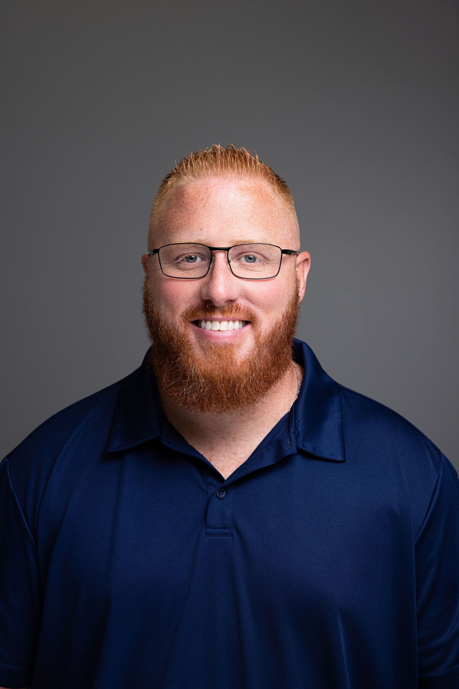Smiling man with red beard and glasses wearing a navy blue polo shirt against a gray background.