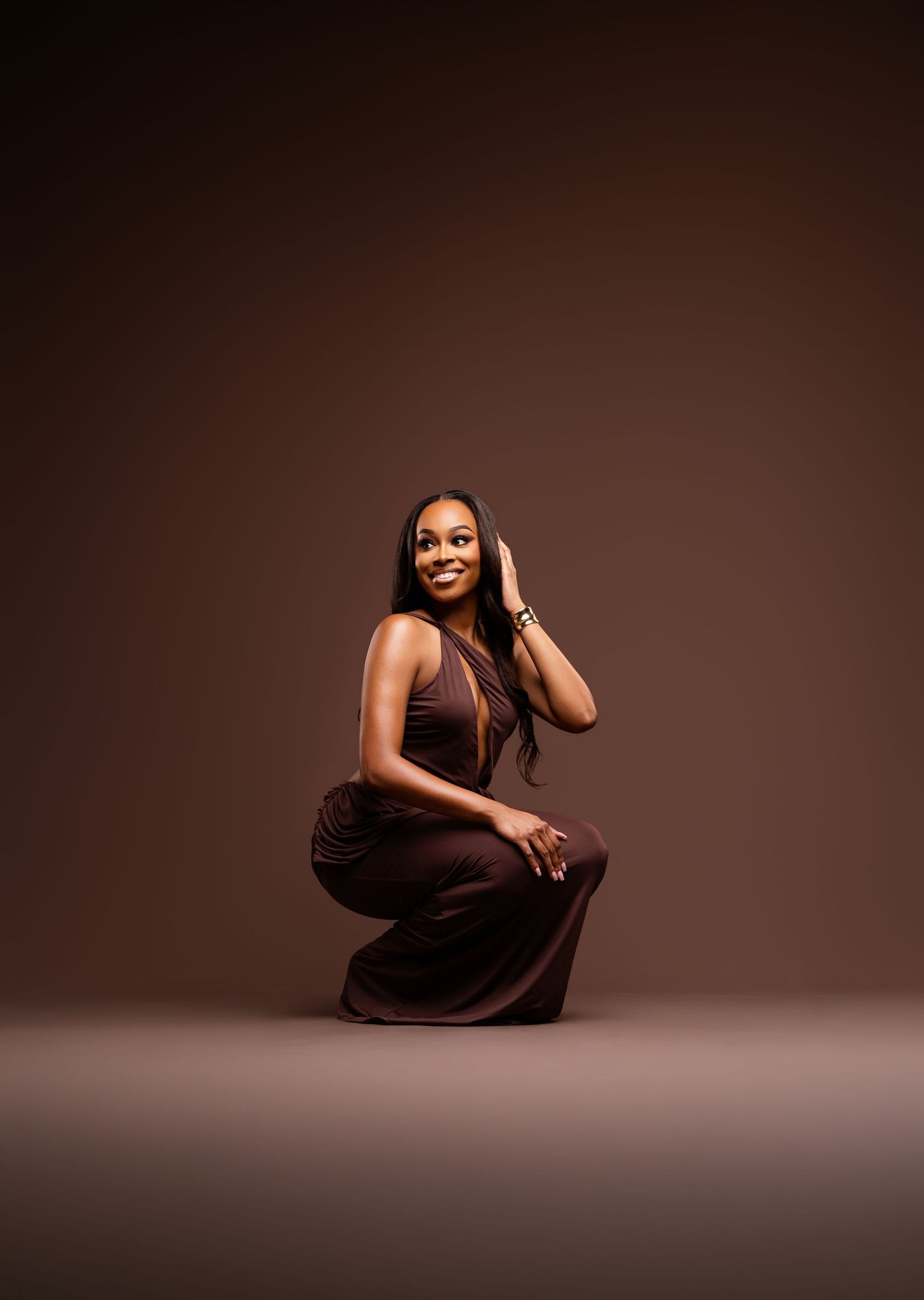 Woman in brown halter jumpsuit posing gracefully on studio floor against warm brown background.