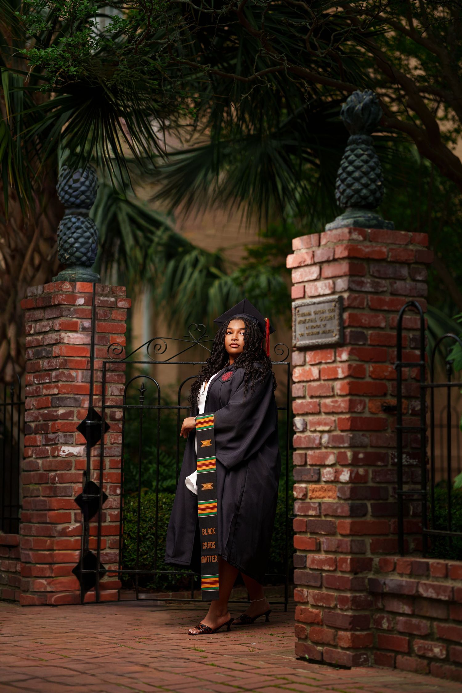 Graduate in black cap and gown with kente stole stands at brick gate surrounded by lush greenery.