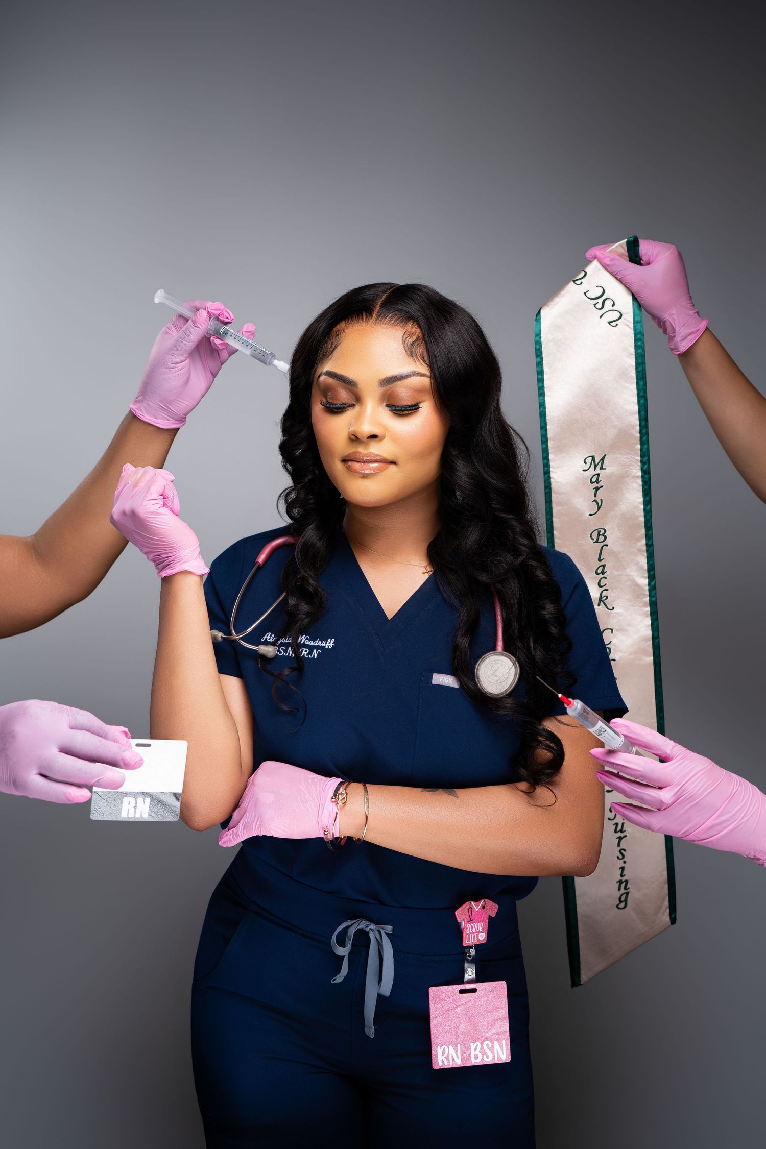 A nurse in blue scrubs holds a graduation sash, surrounded by hands in pink gloves offering syringes and medical supplies.