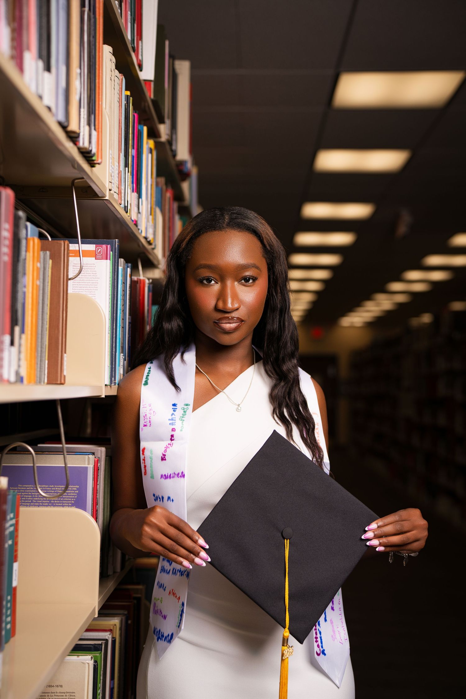 Young Black woman in white dress holds graduation cap while standing in library aisle, celebrating academic achievement.