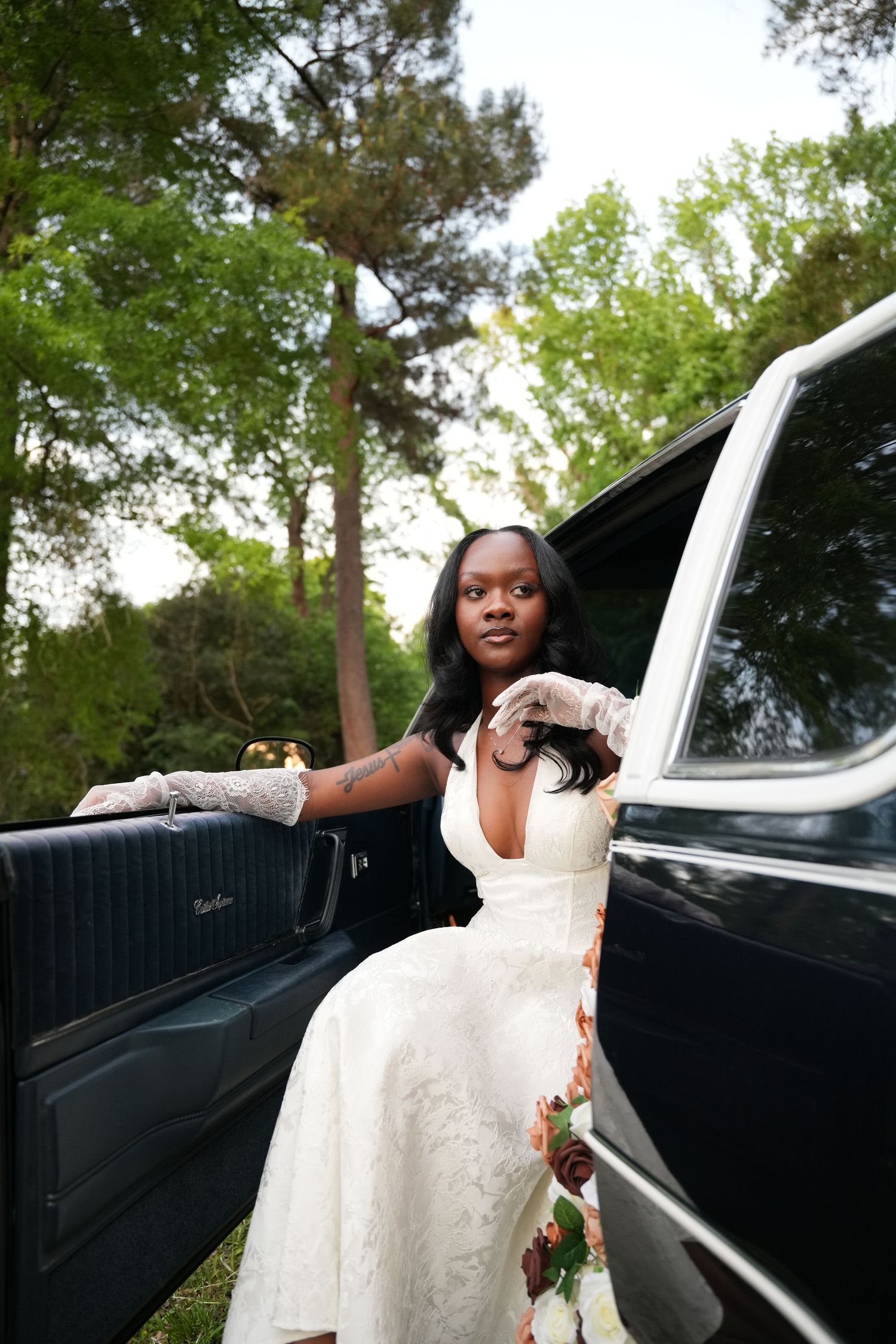 A Black bride in a white floral gown and lace gloves poses beside a vintage black car surrounded by lush green trees.