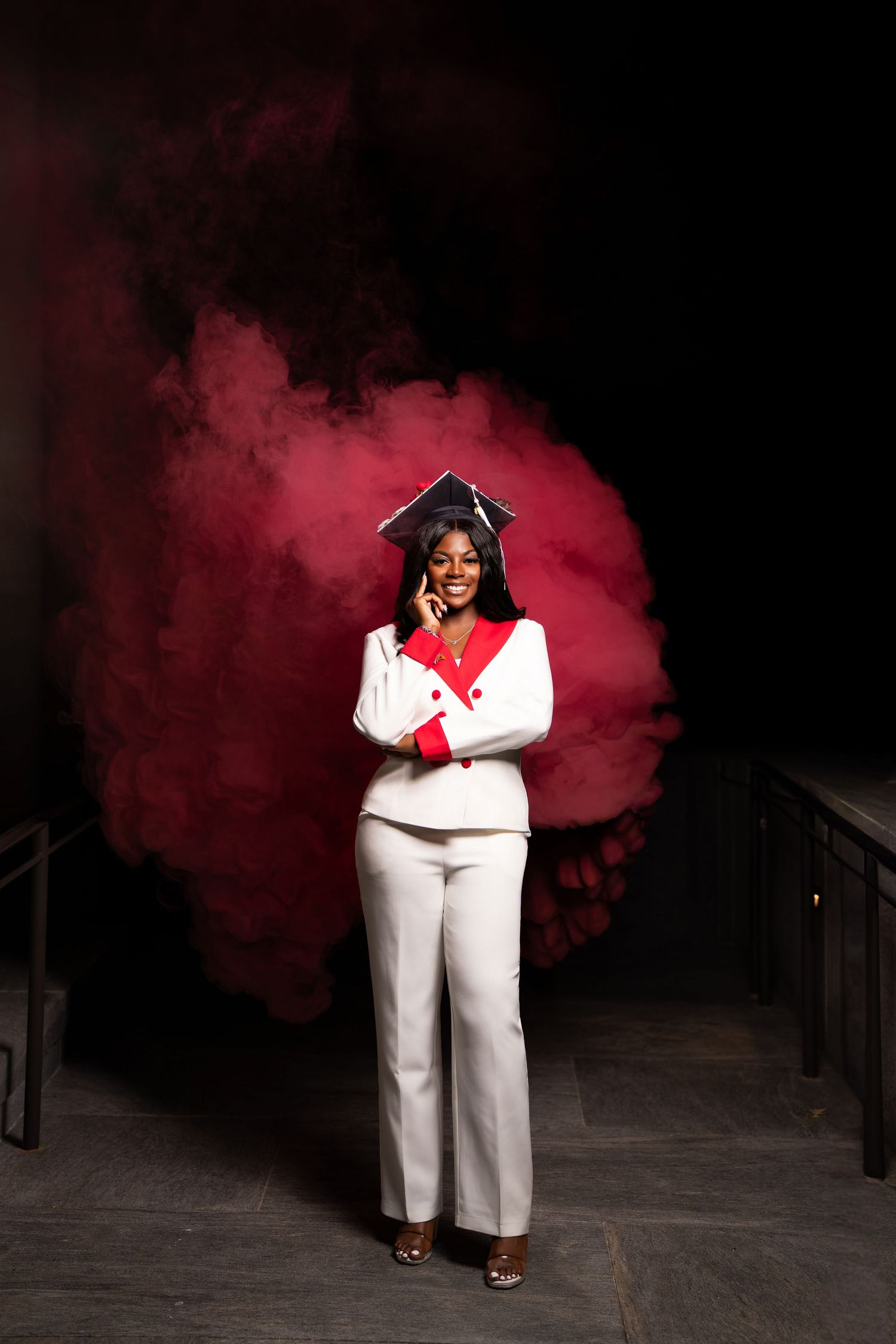 A smiling woman in white suit and graduation cap poses against dramatic red smoke background.