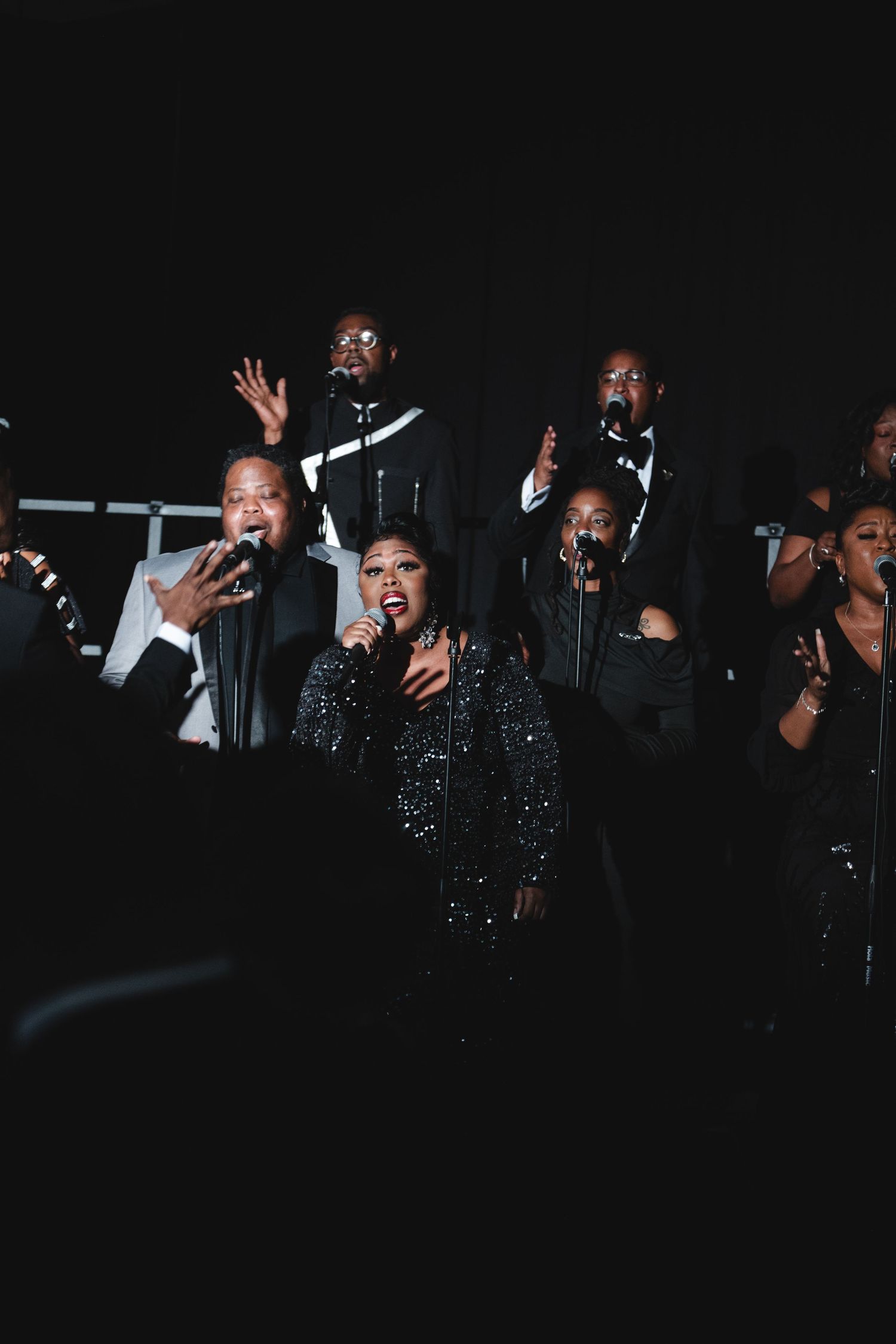Performers in sparkling outfits singing and gesturing dramatically on a dark stage during a show.