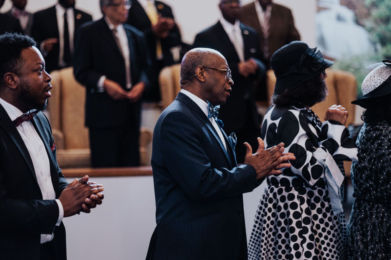 People in formal attire gather at an indoor event, engaged in animated conversation and greetings.