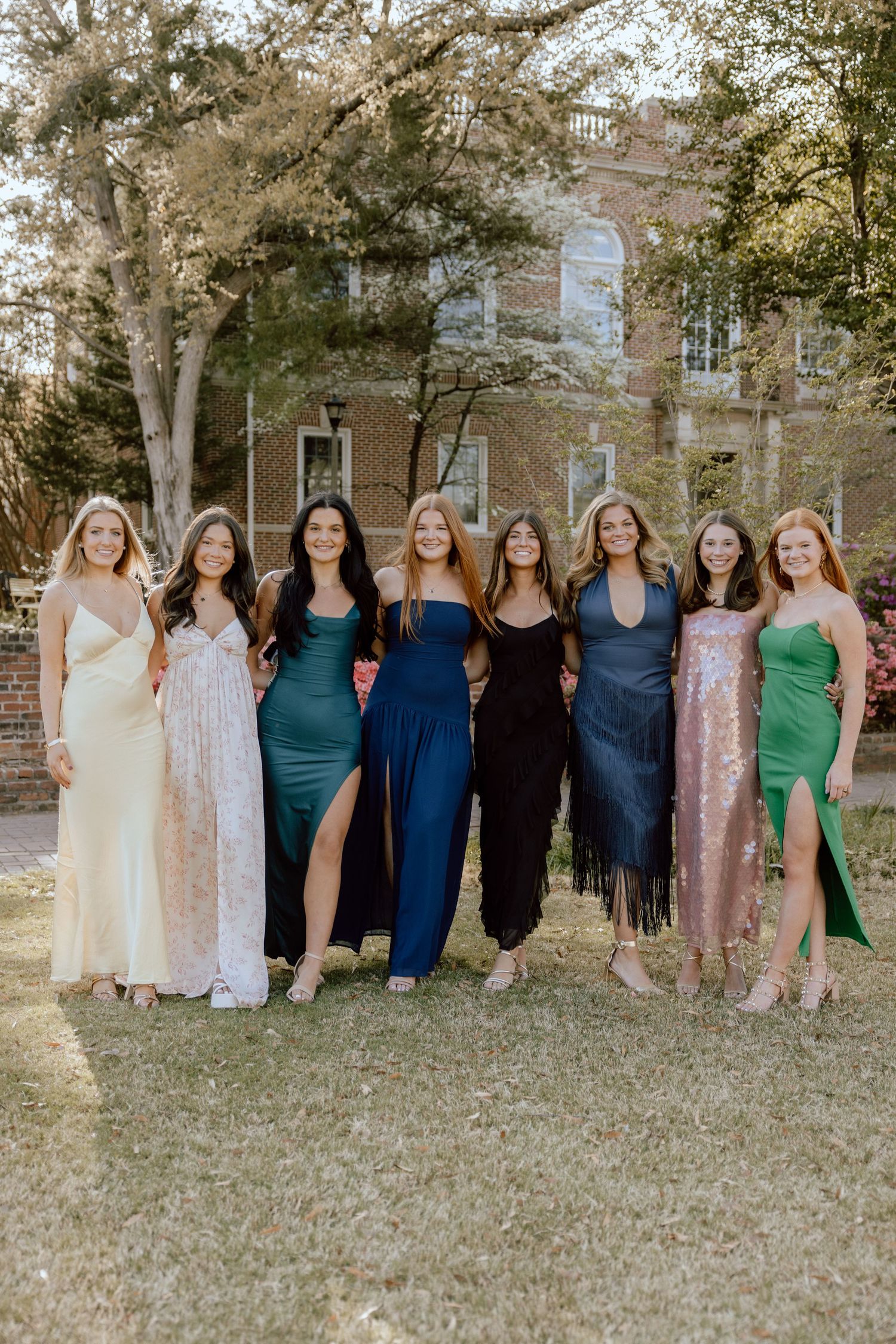 A group of women stand together outdoors wearing different colored dresses in shades of blue, green and cream.