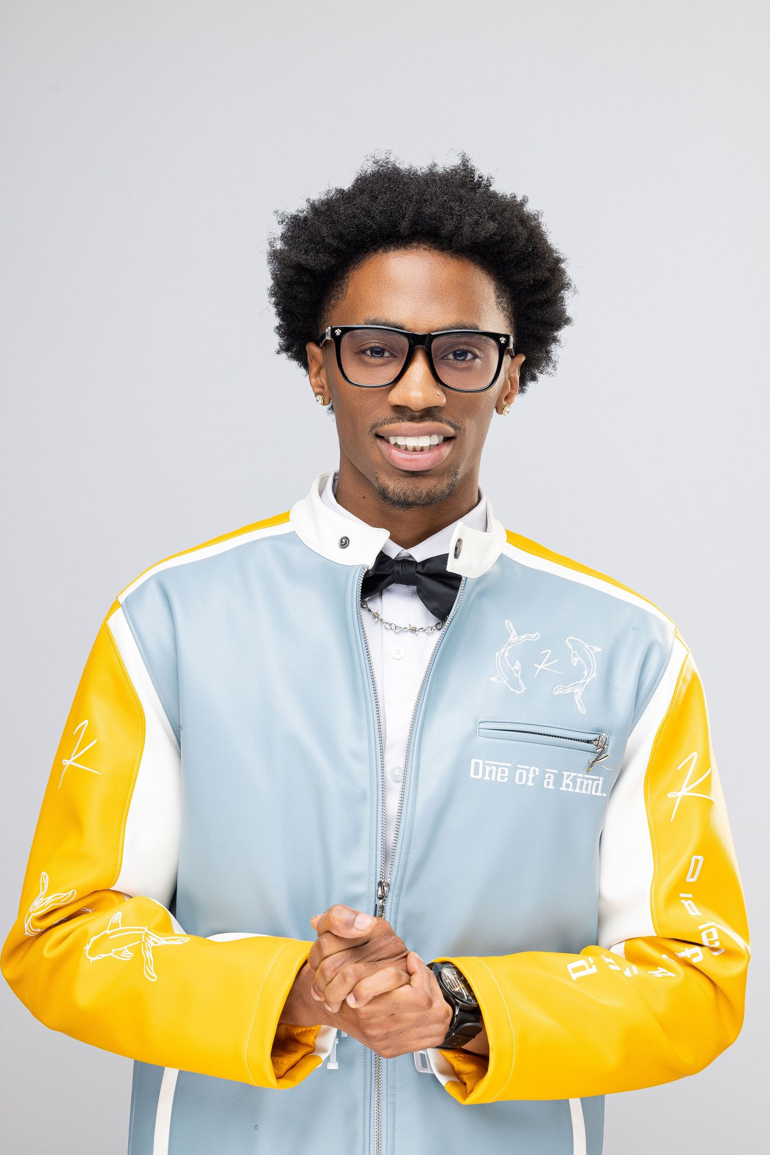 Person wearing glasses and bow tie in a stylish light blue and yellow varsity jacket smiles at camera.