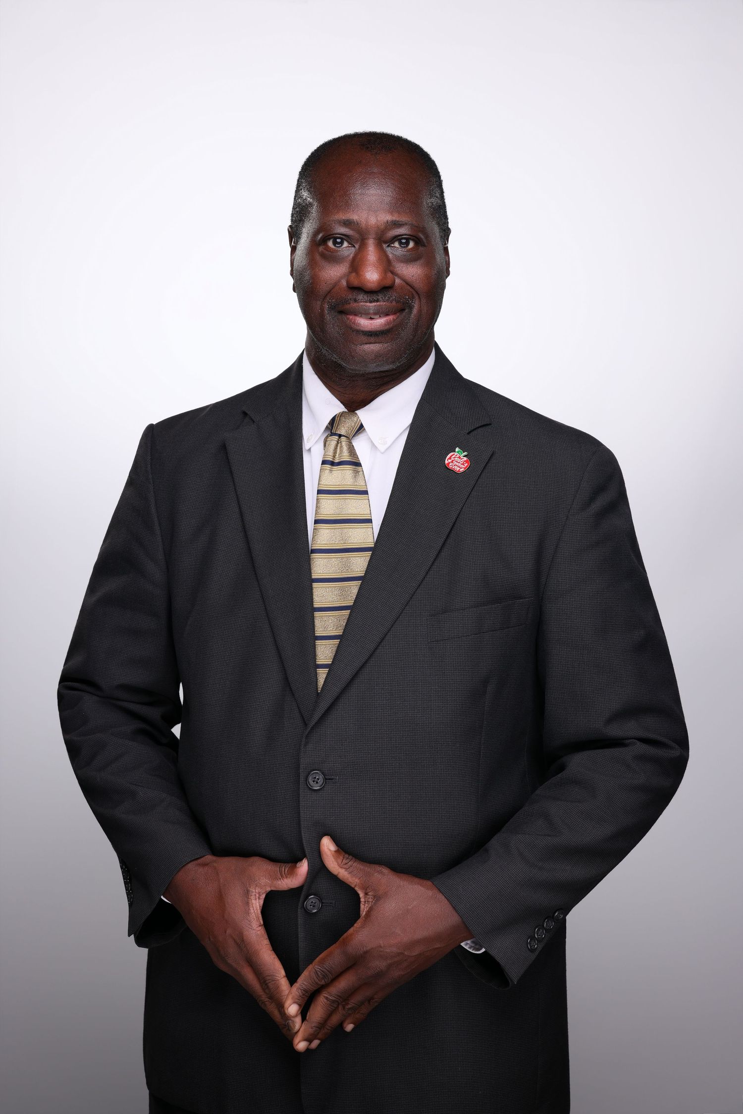 Professional man in black suit with striped tie and lapel pin, smiling confidently against a light gray background.