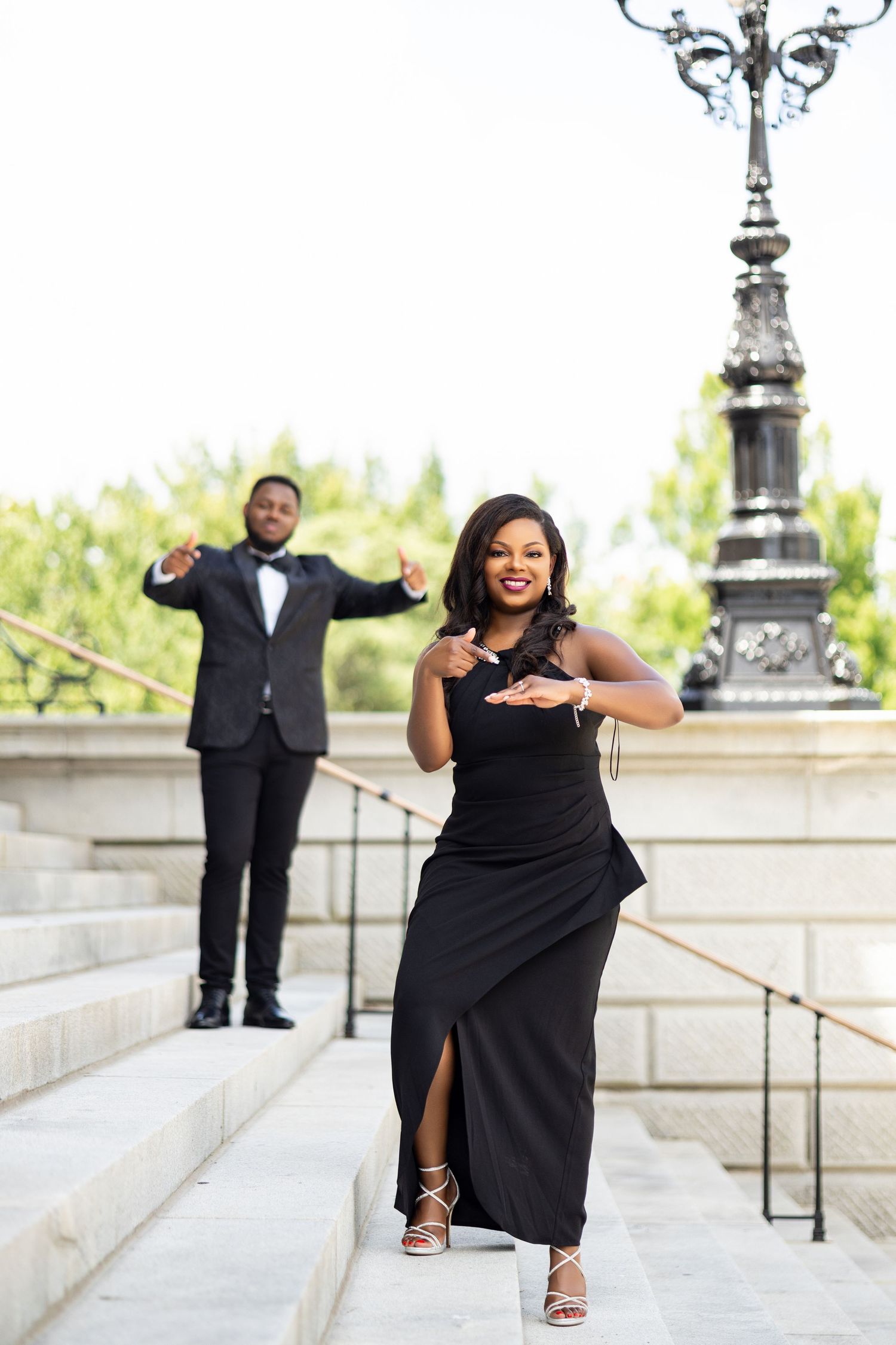 Couple in formal wear strike dramatic pose on outdoor steps near ornate lamppost.