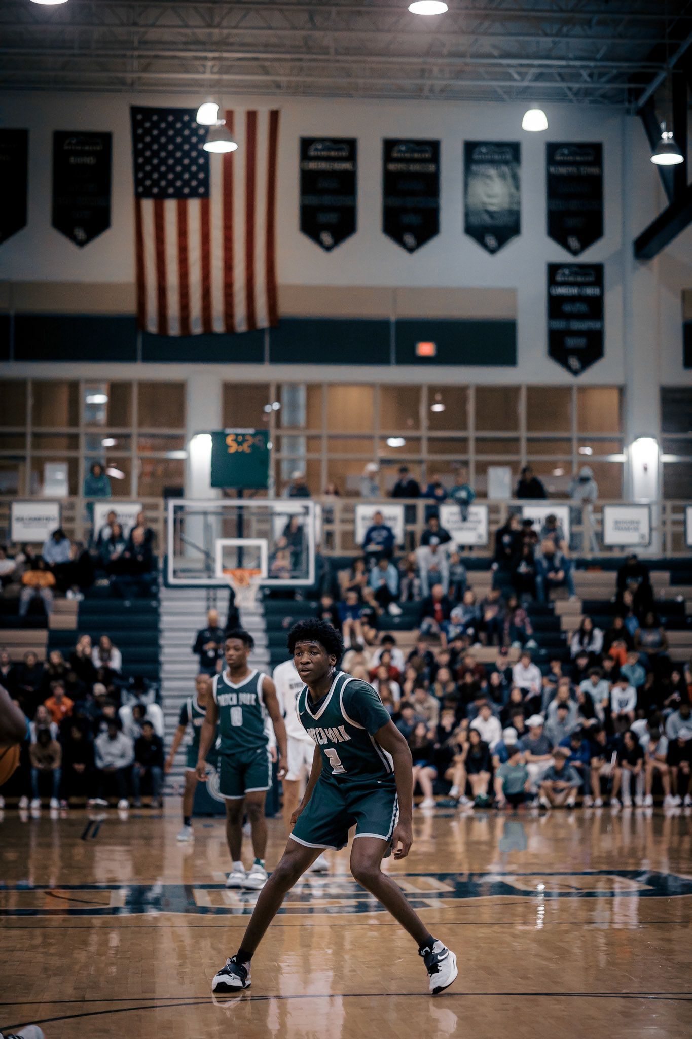 Basketball game in progress with American flag and championship banners displayed in gymnasium.
