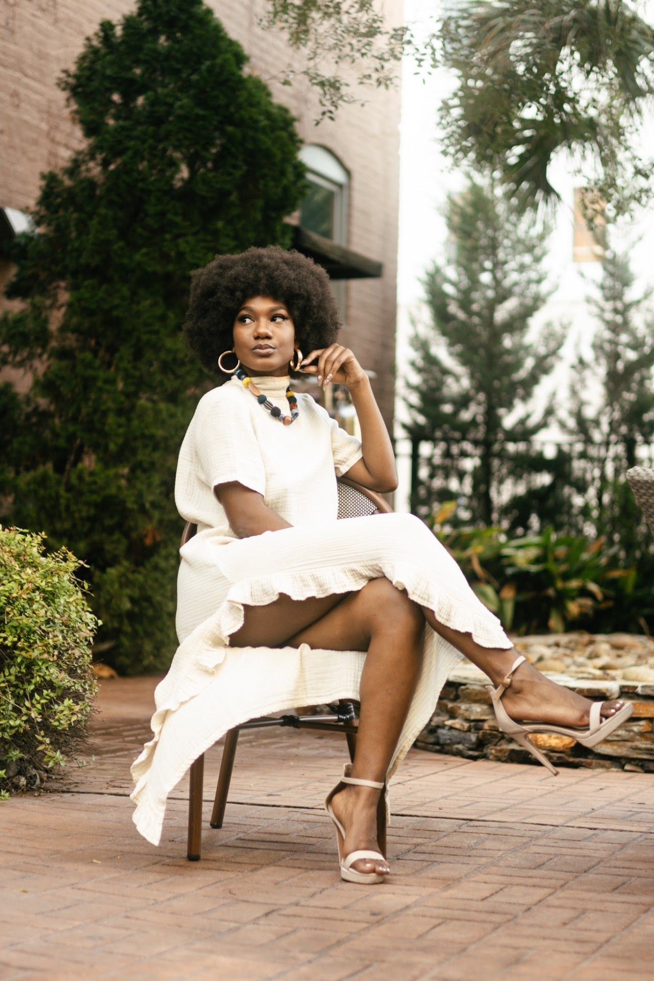 A model in a flowing white dress and sandals poses elegantly on a chair on an outdoor patio surrounded by greenery.
