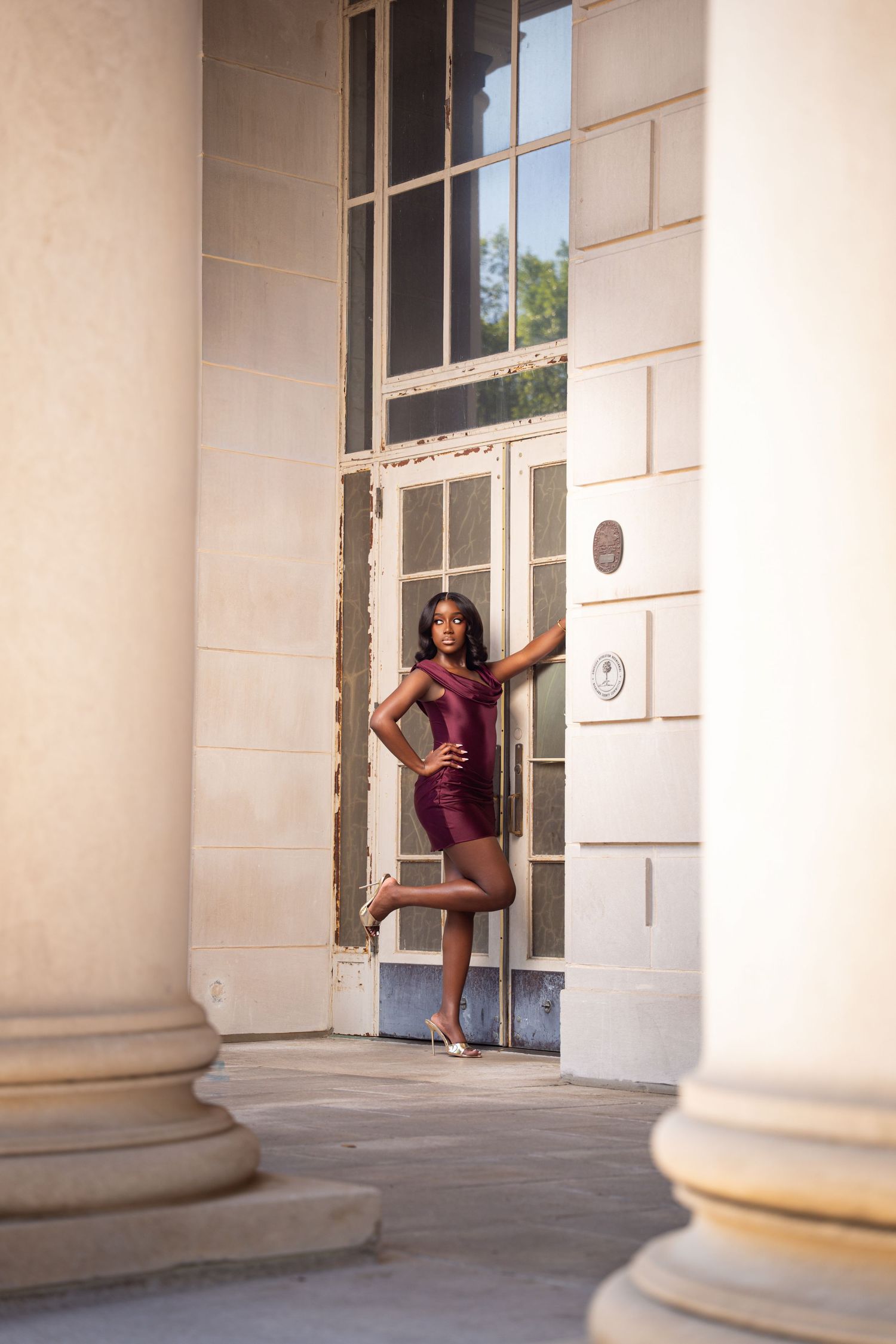 A woman in a burgundy mini dress poses gracefully between classical columns of an elegant building.