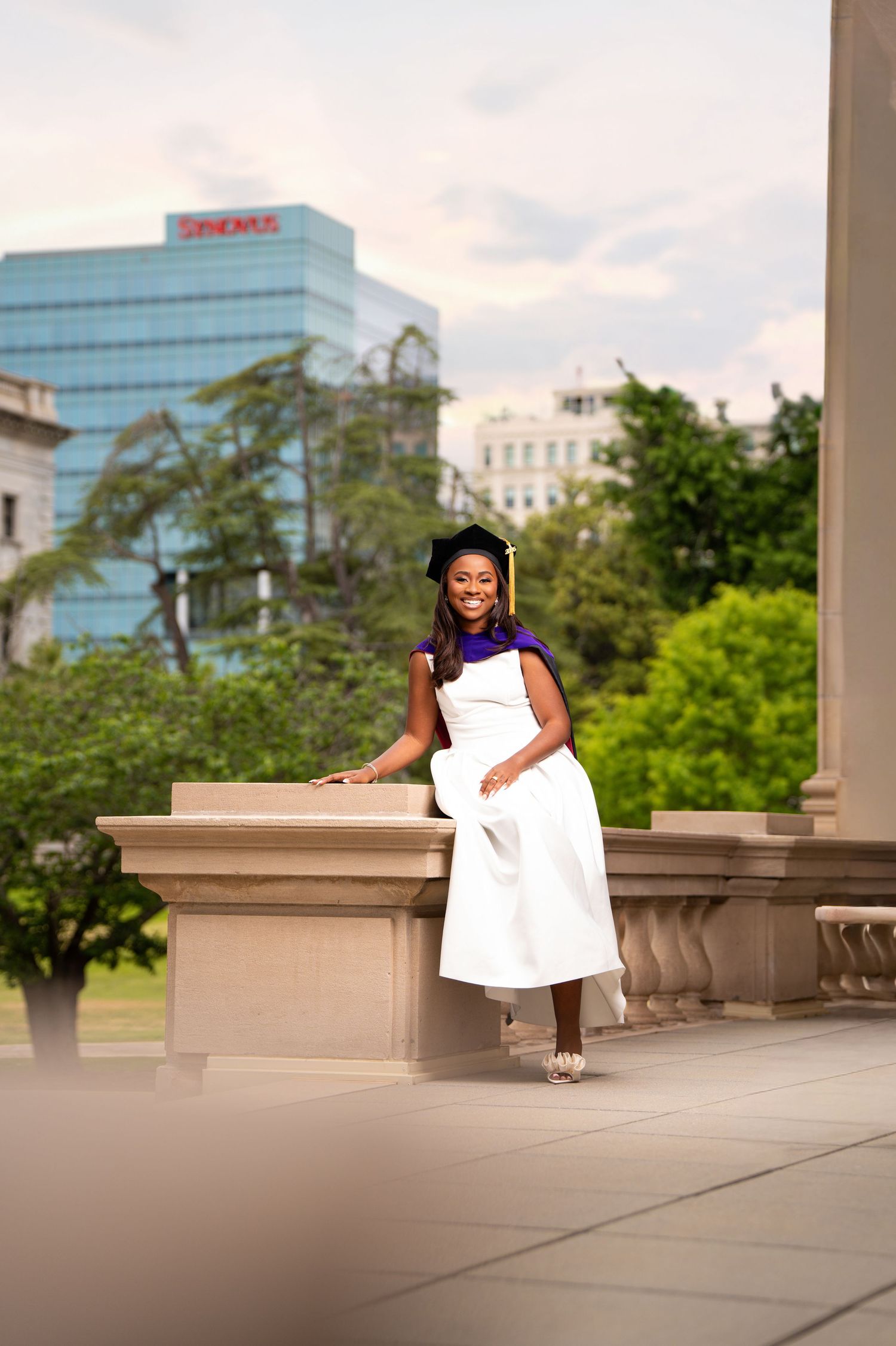 Smiling graduate in white dress and cap poses on stone columns with green trees and city buildings in background.