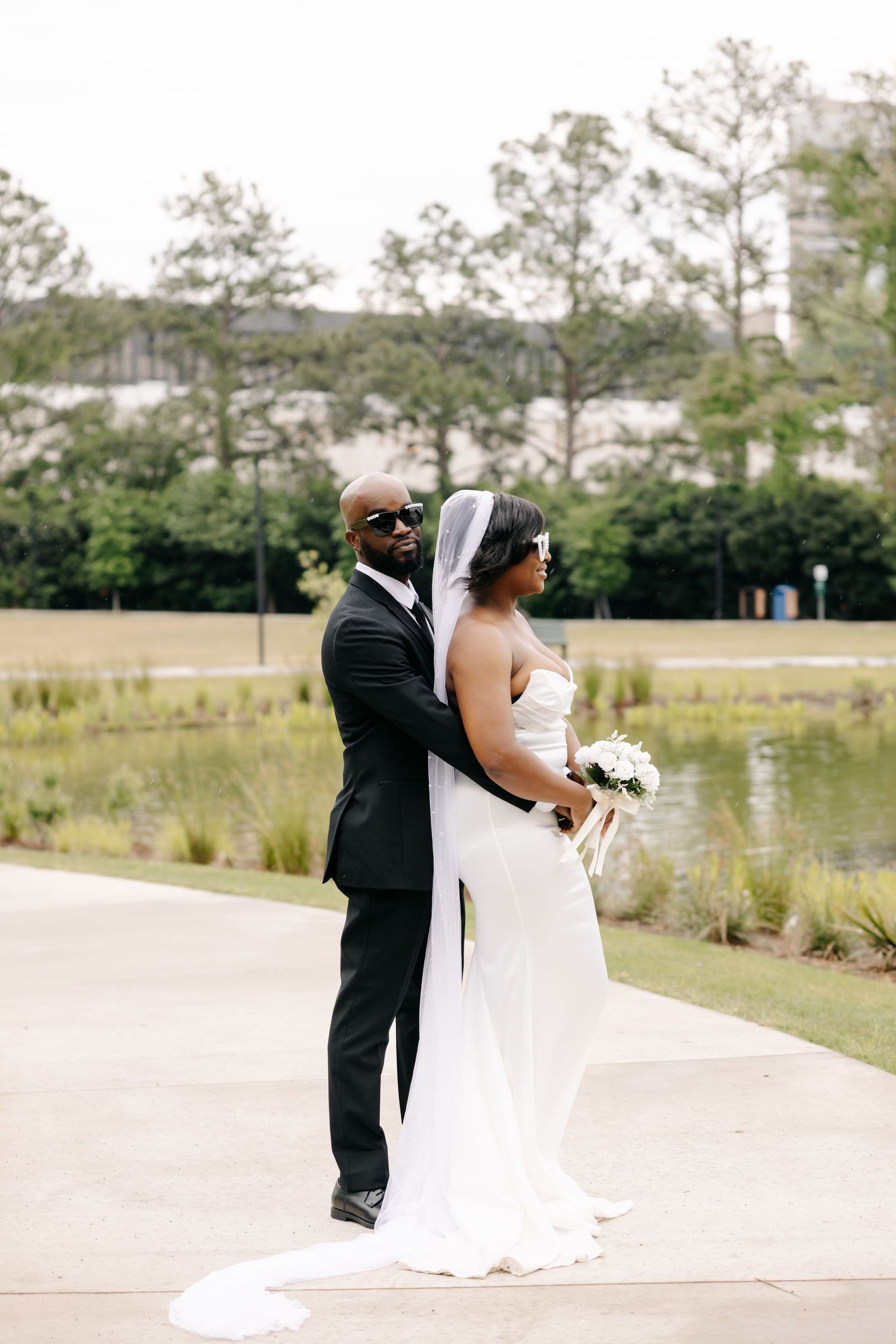 A bride and groom pose elegantly outdoors near a pond, she in a white gown with veil, he in a black tuxedo with sunglasses.