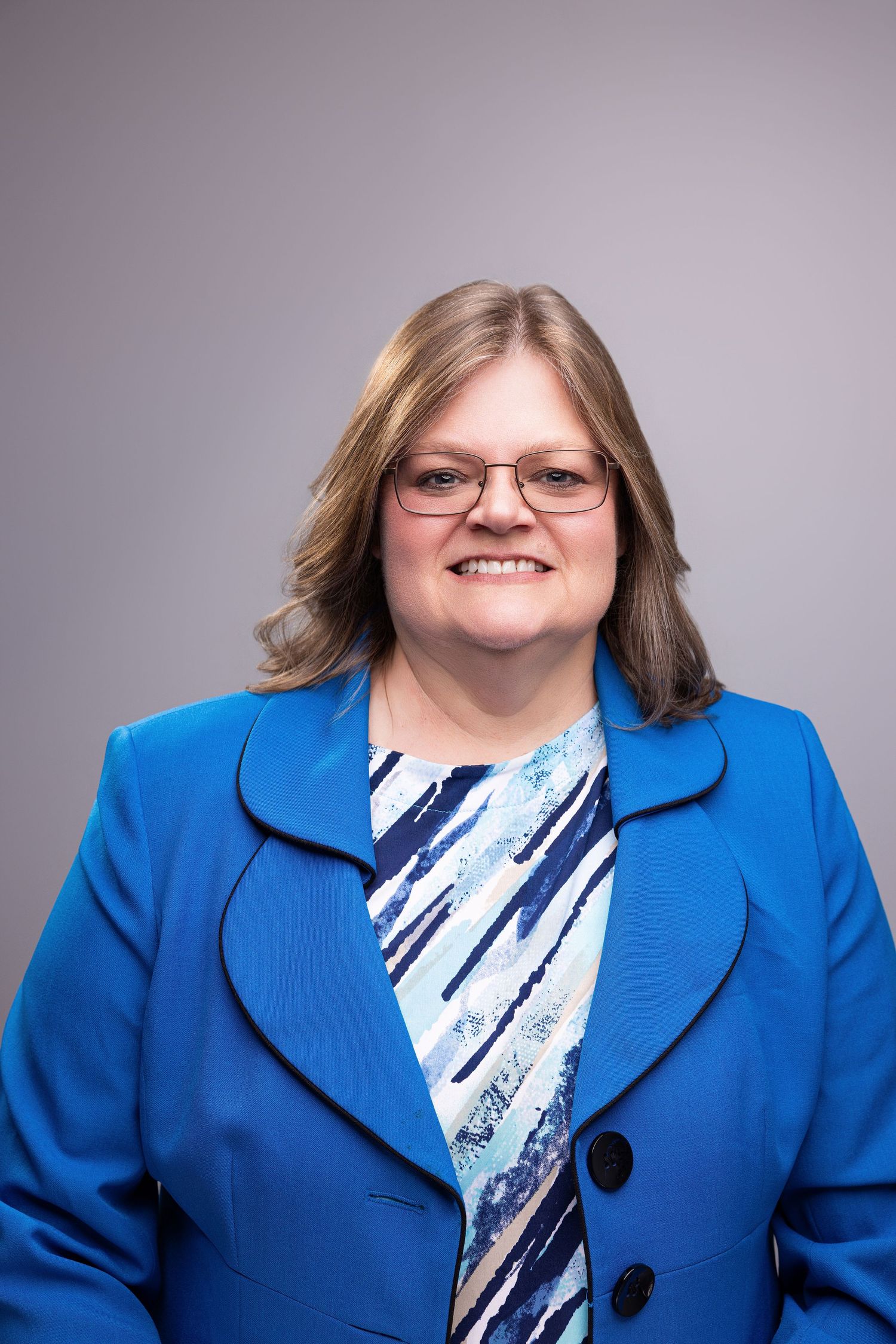 Smiling woman in blue blazer and glasses poses for professional headshot against gray background.