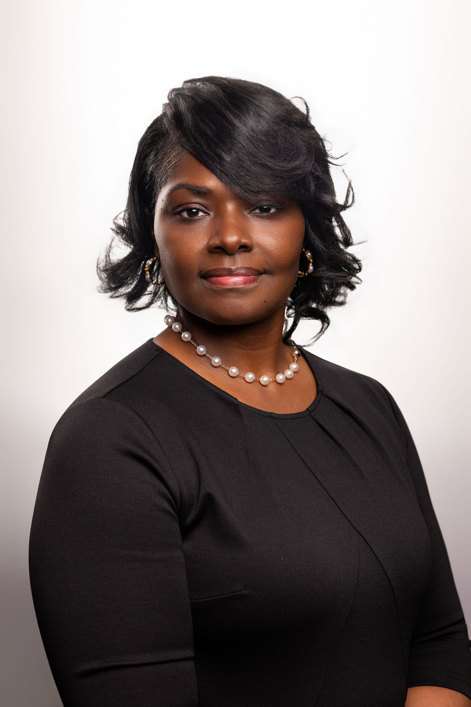 Professional headshot of a woman in a black dress wearing pearl necklace and earrings, posing against a light background.