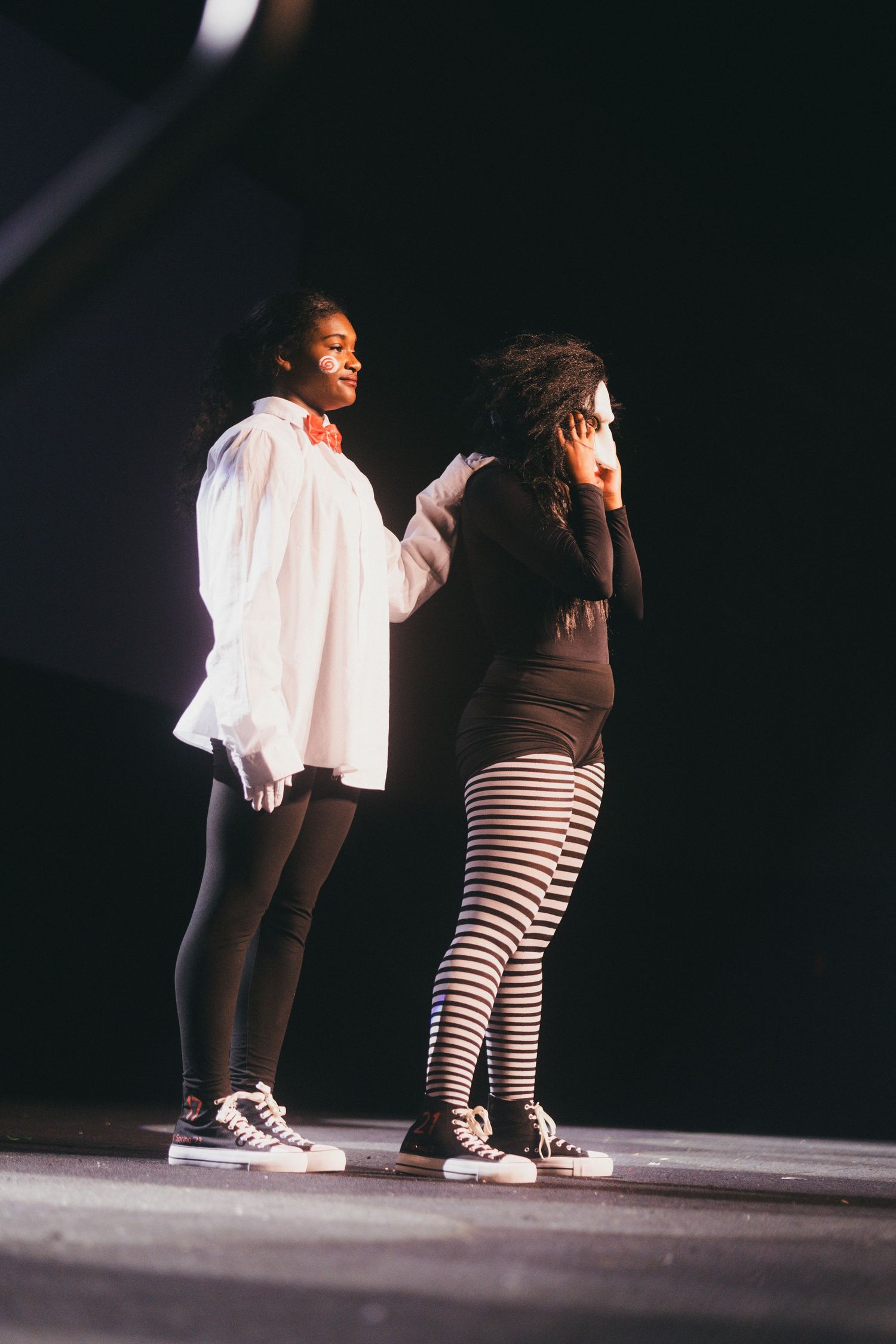 Two deltas  on stage, one wearing a white shirt and dark pants, another in striped leggings and black top in dramatic lighting.