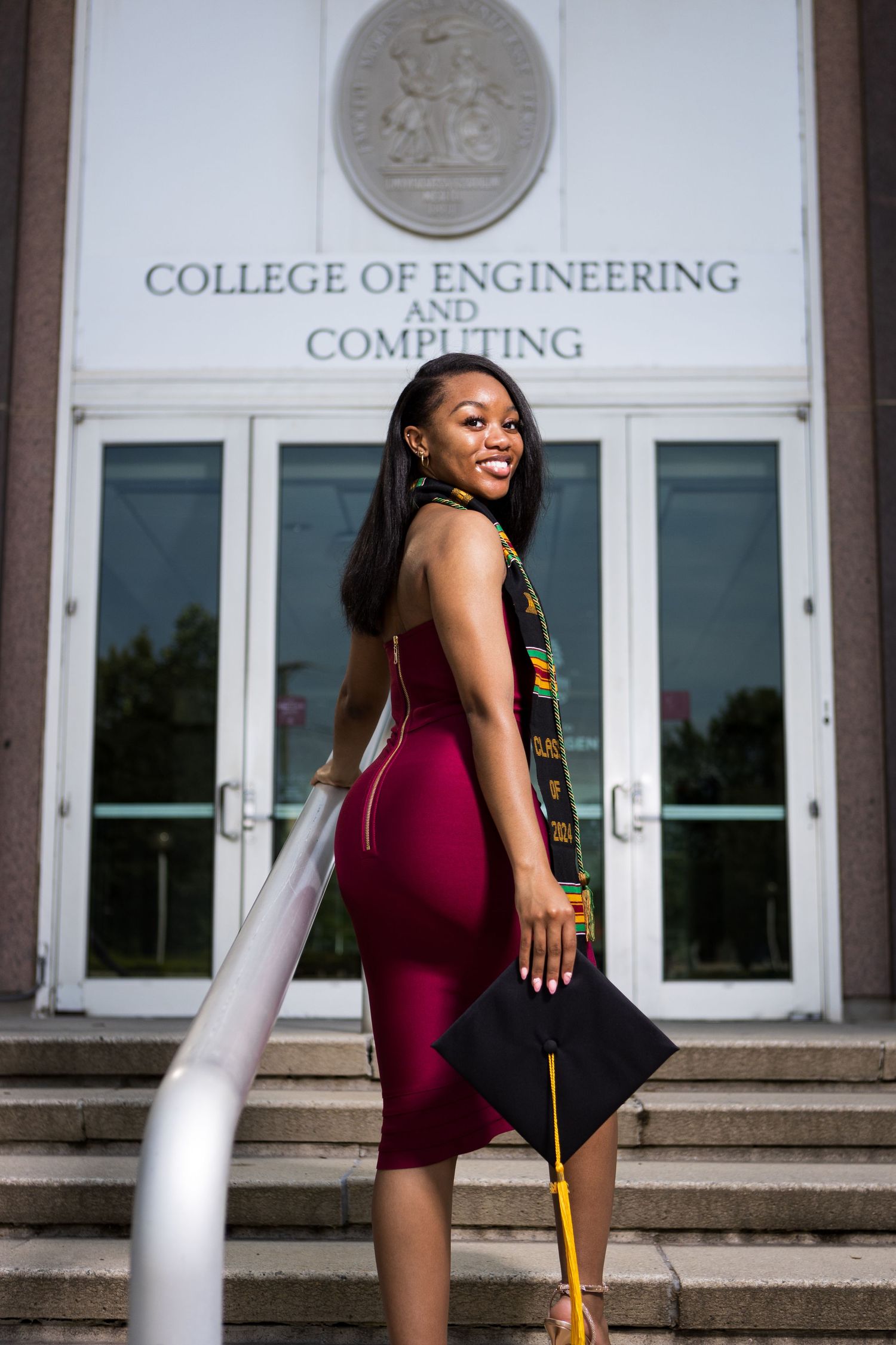 A graduate in a burgundy dress poses confidently on steps outside the College of Engineering building.