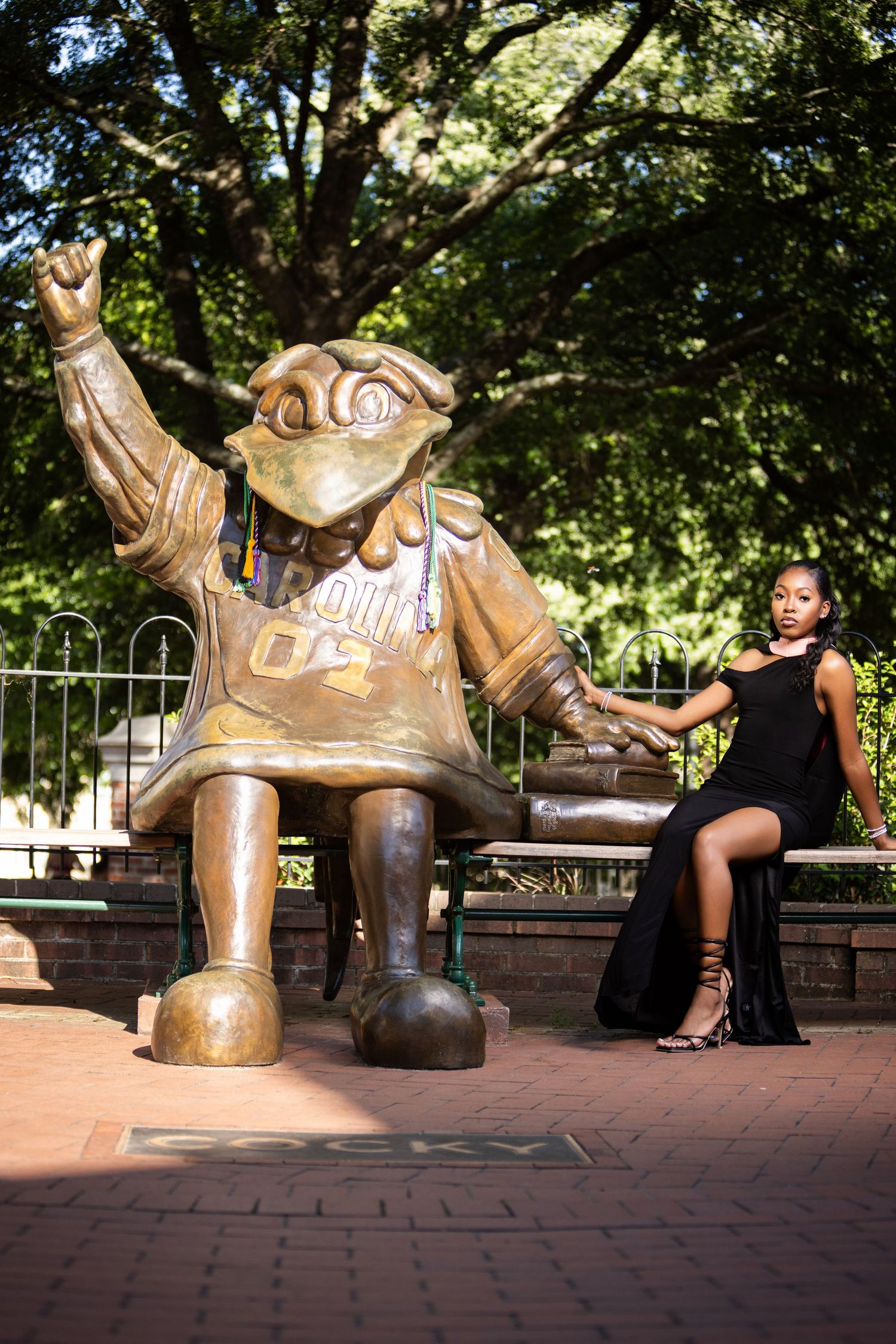 Person in black dress posing next to large bronze mascot statue in outdoor campus setting.