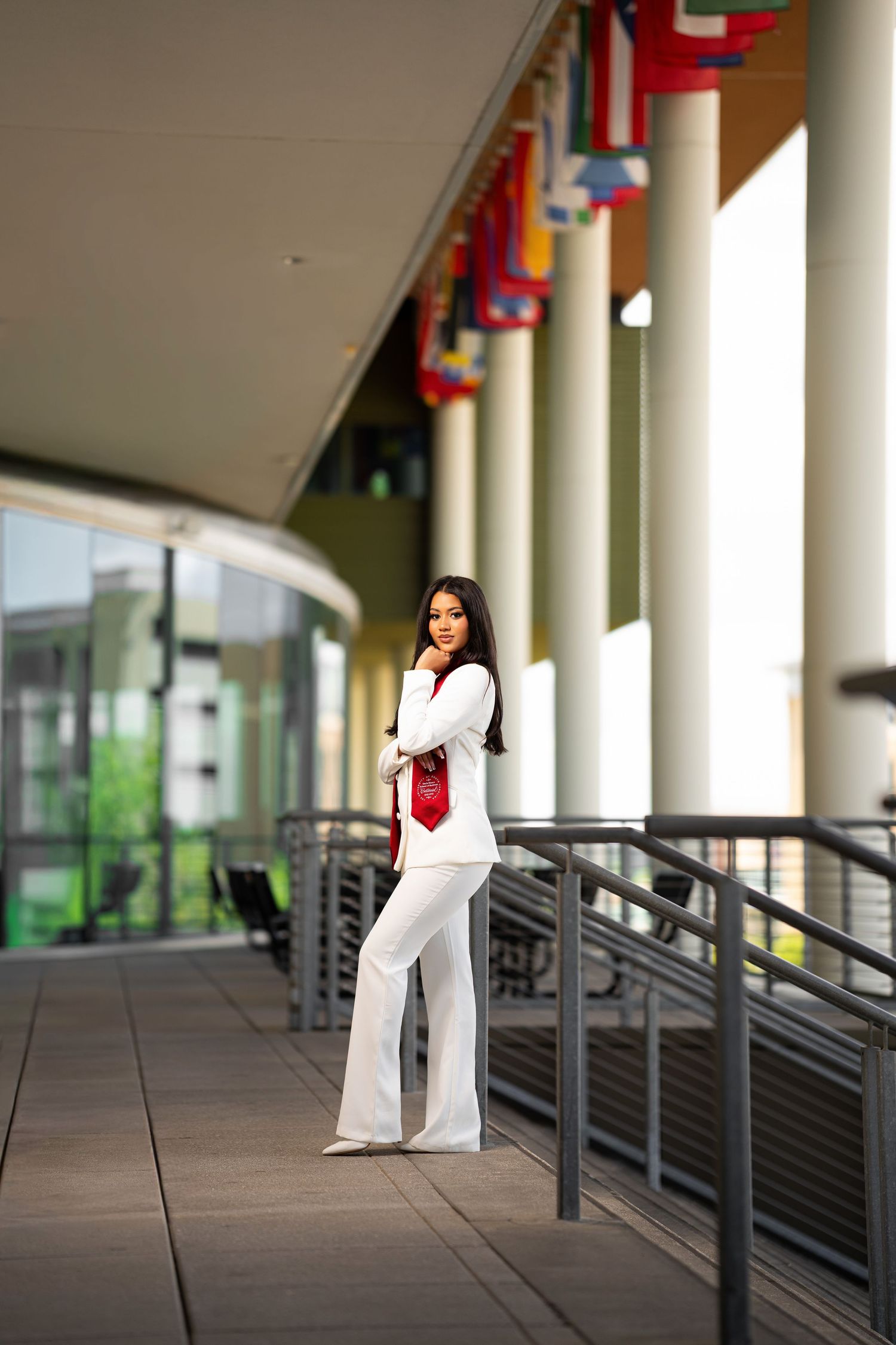Young woman in white pantsuit with red tie poses confidently by railing outside building with international flags.