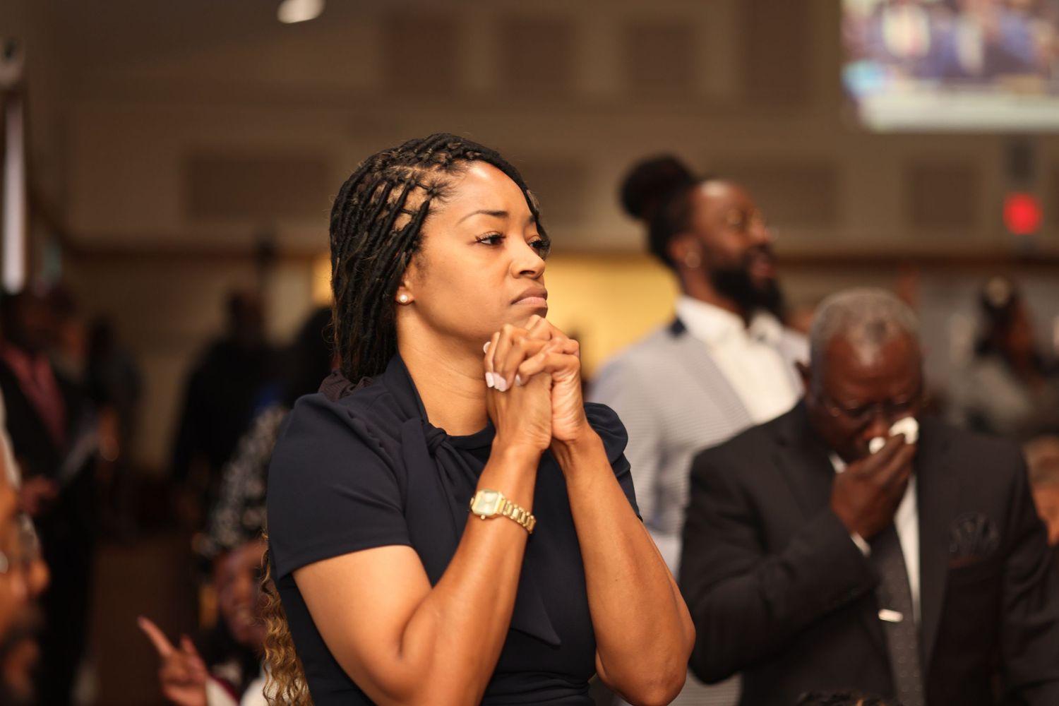 A person in a black shirt clasps their hands together while watching intently in a crowded indoor setting.