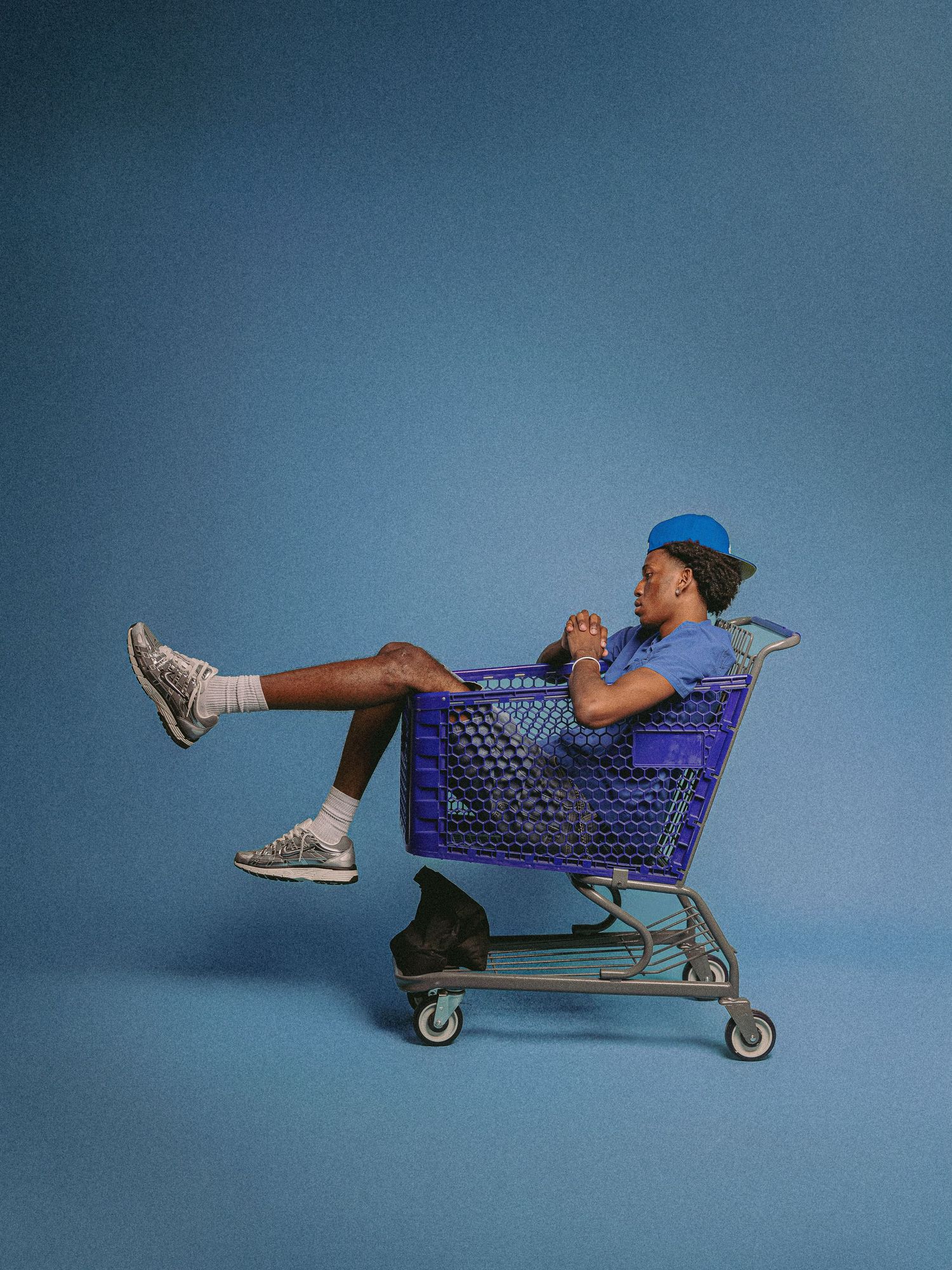 A person lies playfully in a purple shopping cart against a blue background in a creative photoshoot setup.