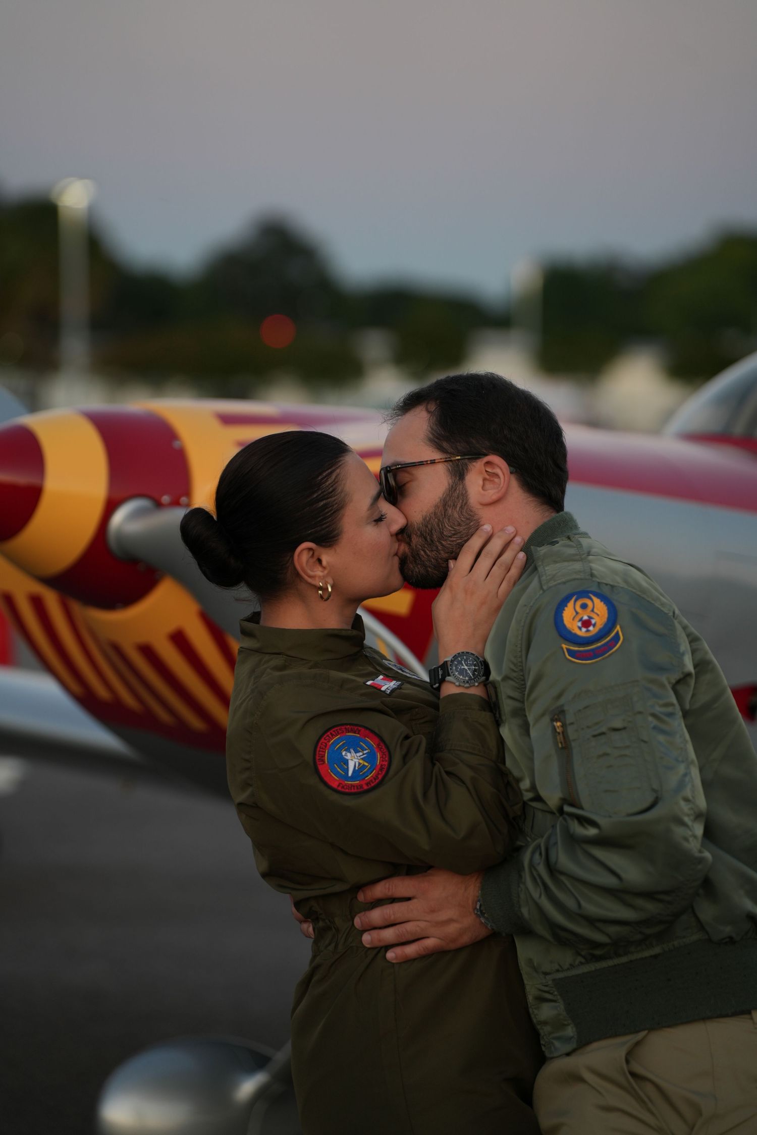 Couple in matching olive green flight suits sharing a kiss on an airport tarmac beside a colorful aircraft.