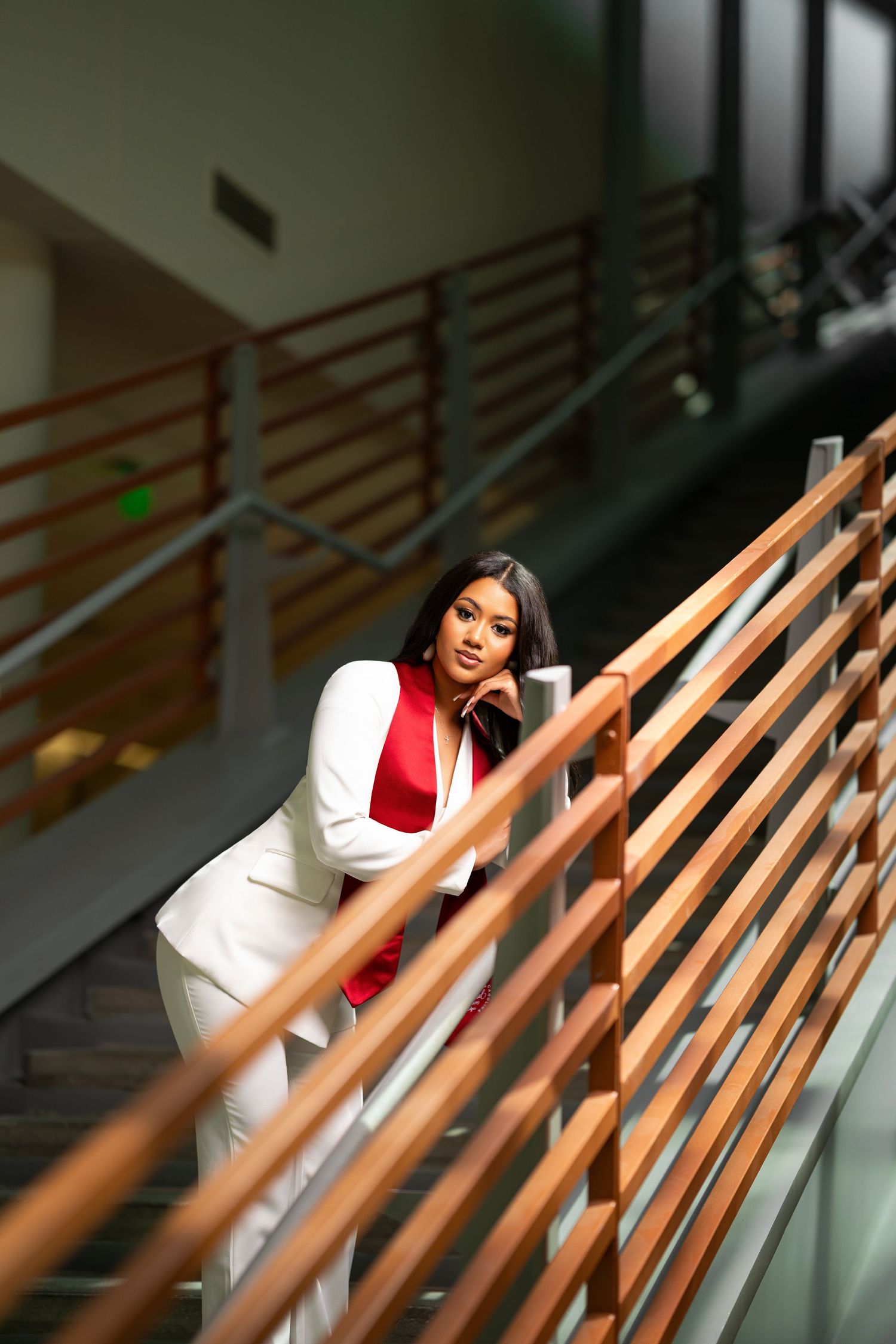 A woman in a white suit with red top leans gracefully on wooden stair railings in a modern building.