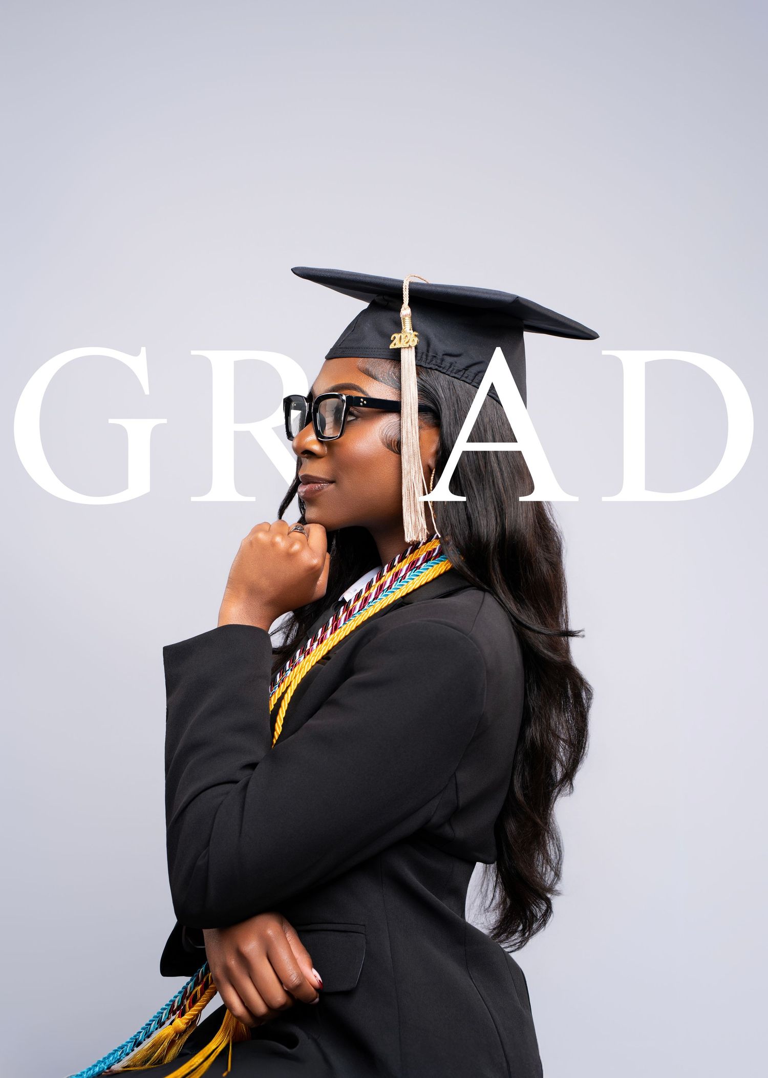 A young Black woman in graduation cap, gown, and colorful honor cords poses thoughtfully against a gray background.