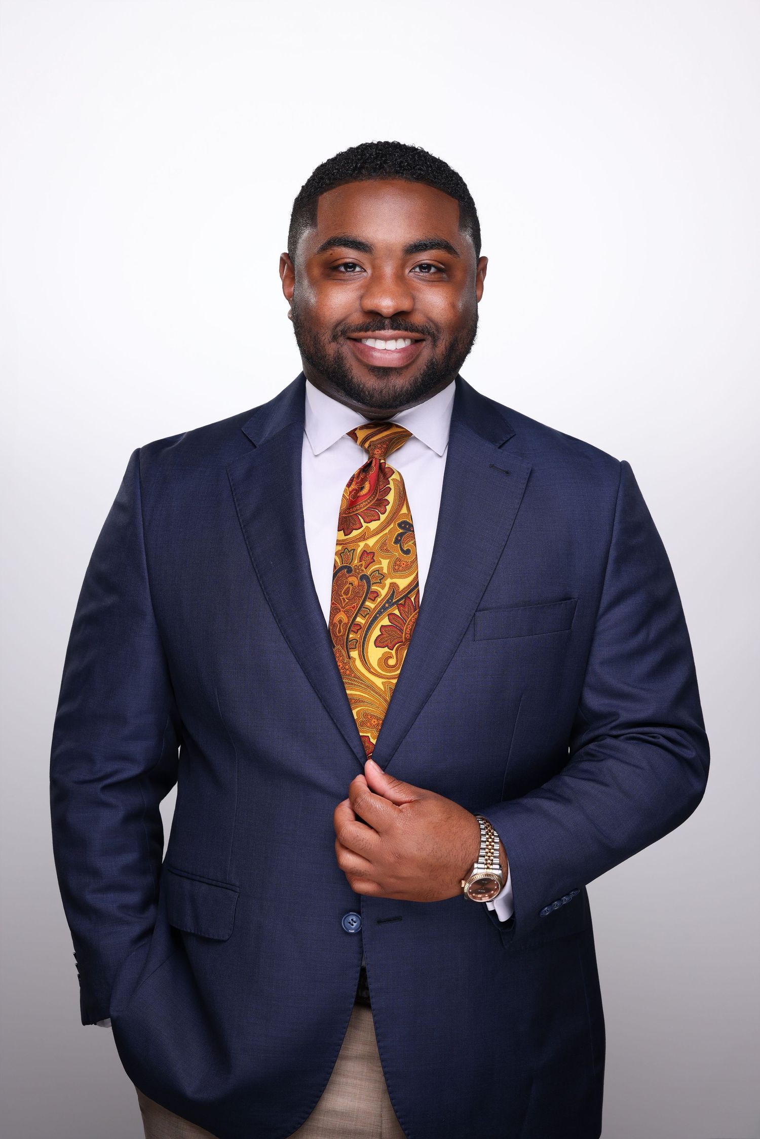 Professional headshot of a smiling young man in a navy suit with a gold paisley tie, holding his lapel confidently.