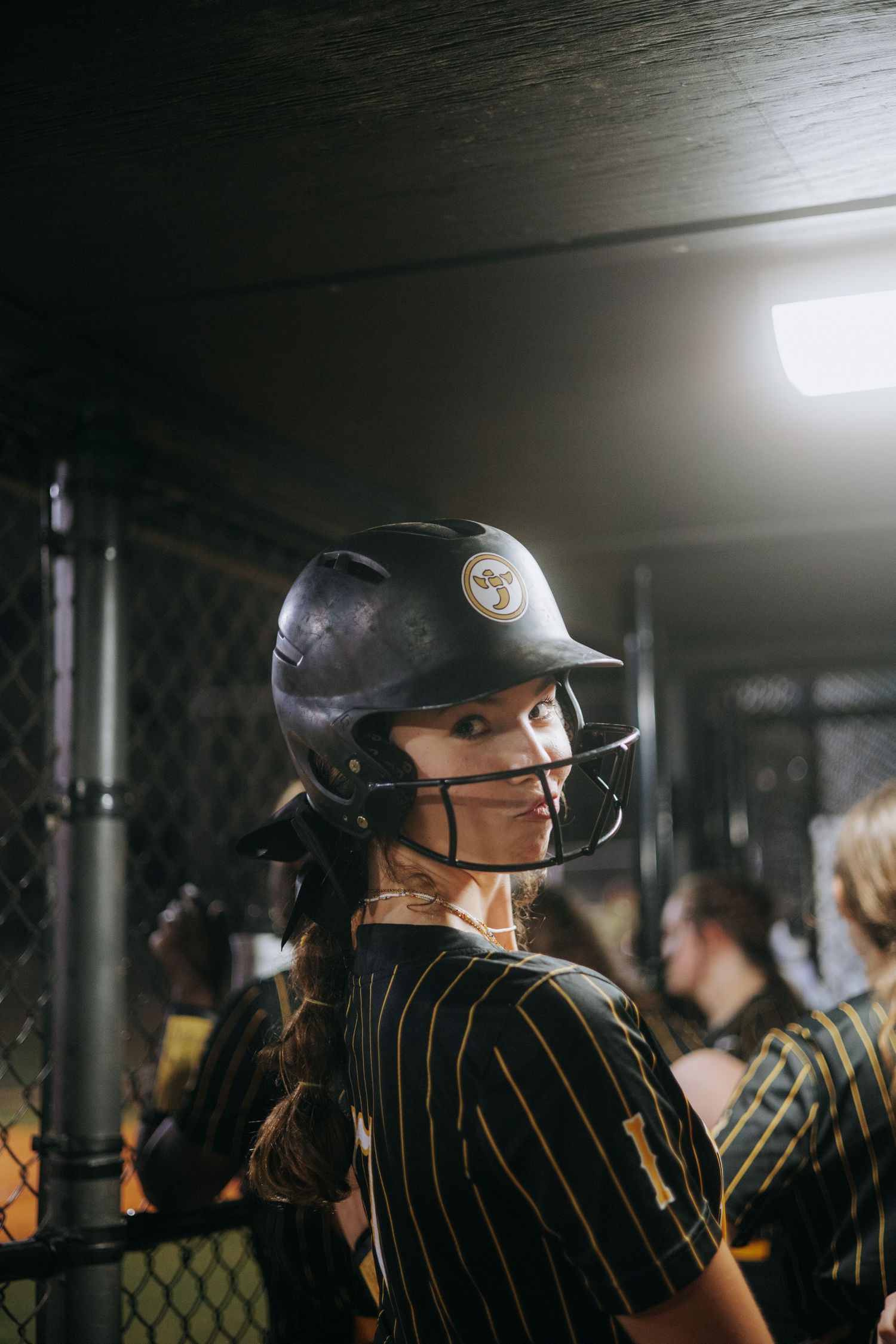Softball player in pinstriped uniform and helmet stands ready in dugout.