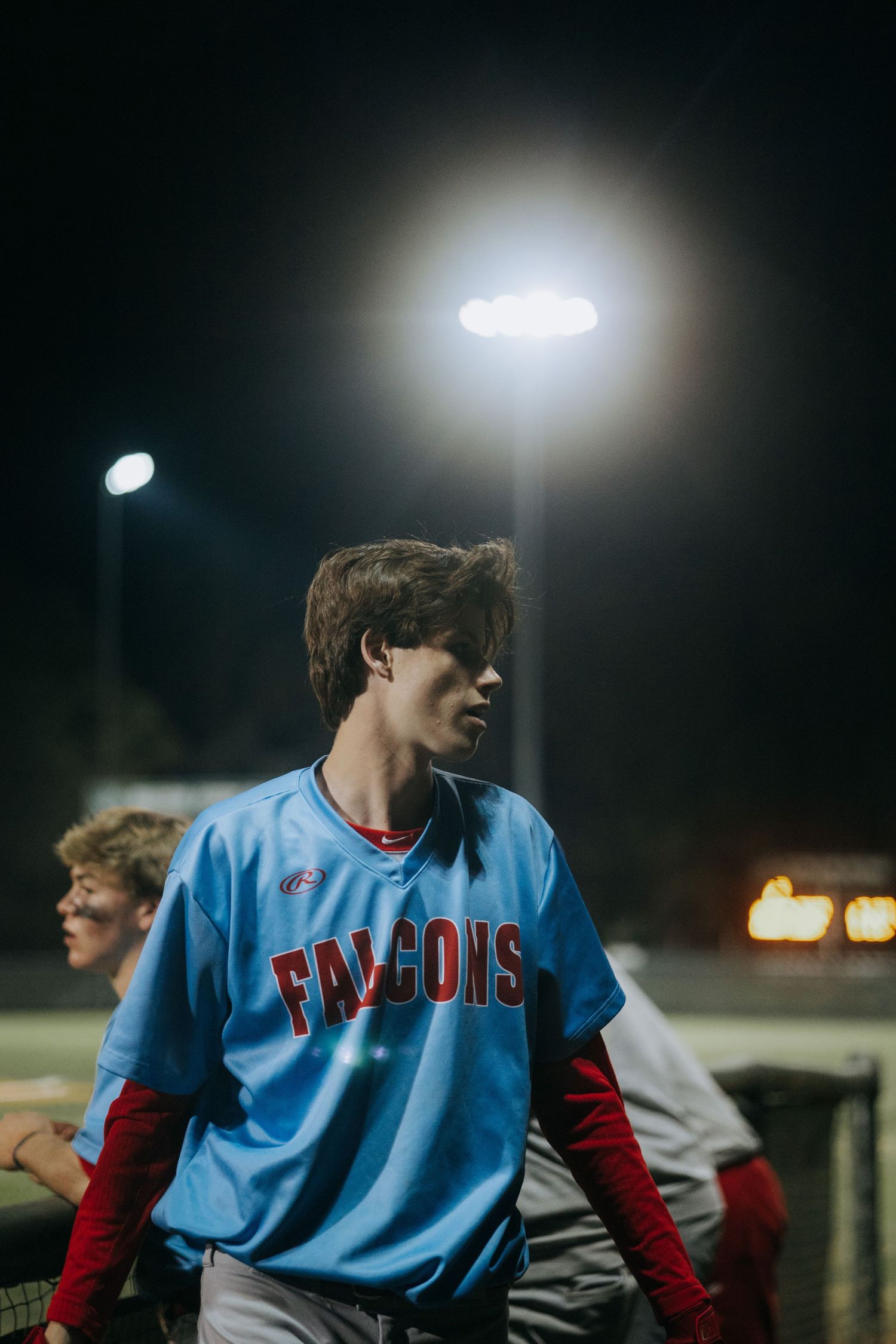High school baseball player in blue Falcons jersey stands in dugout during night game.