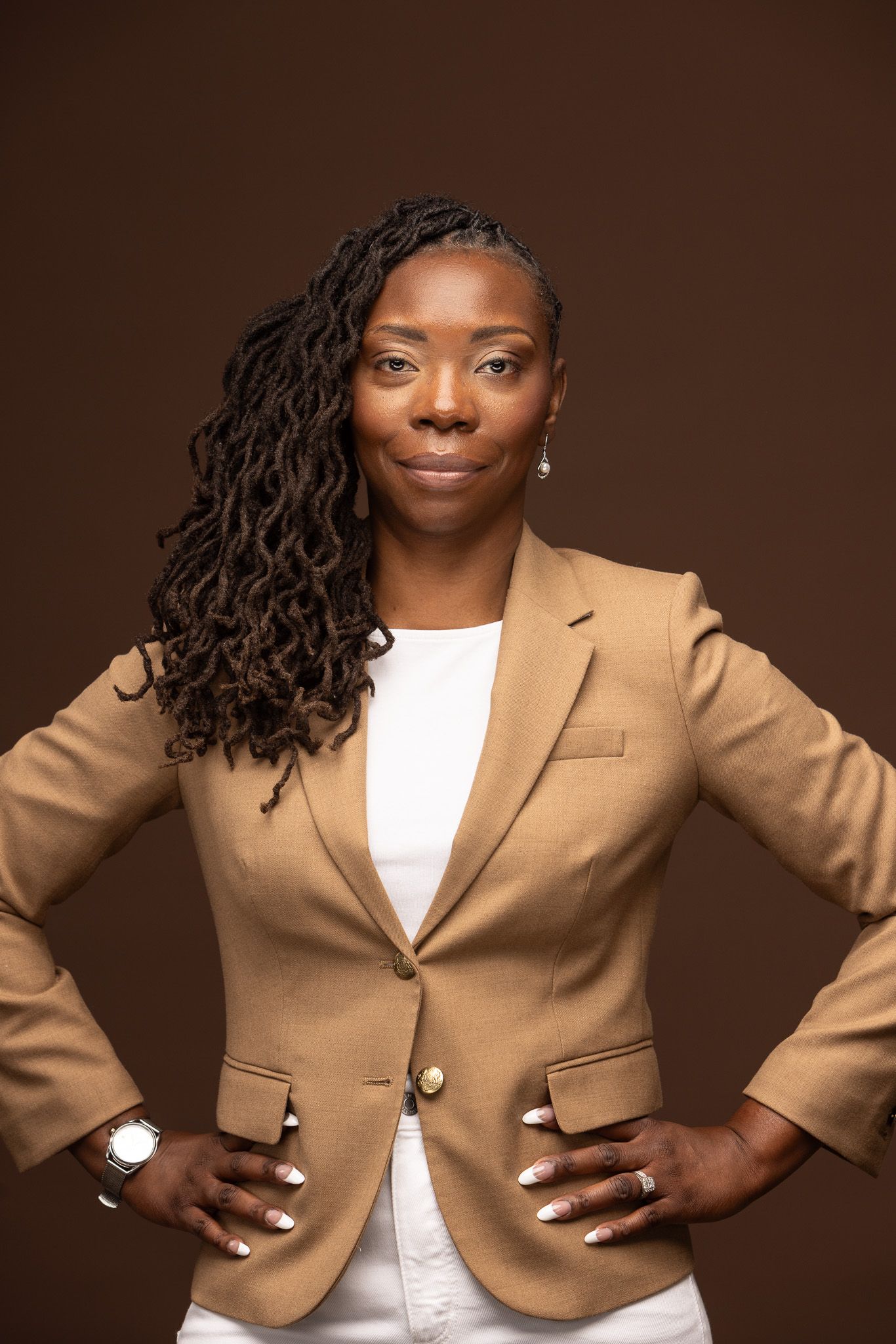 Professional portrait of person in tan blazer and white top with long locs against brown background.