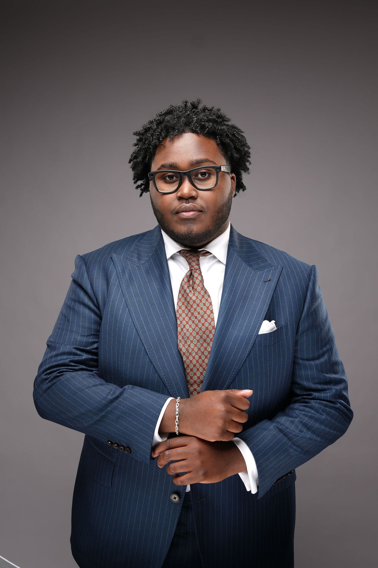 Professional studio portrait of someone in a pinstriped navy suit and brown tie against a gray backdrop.