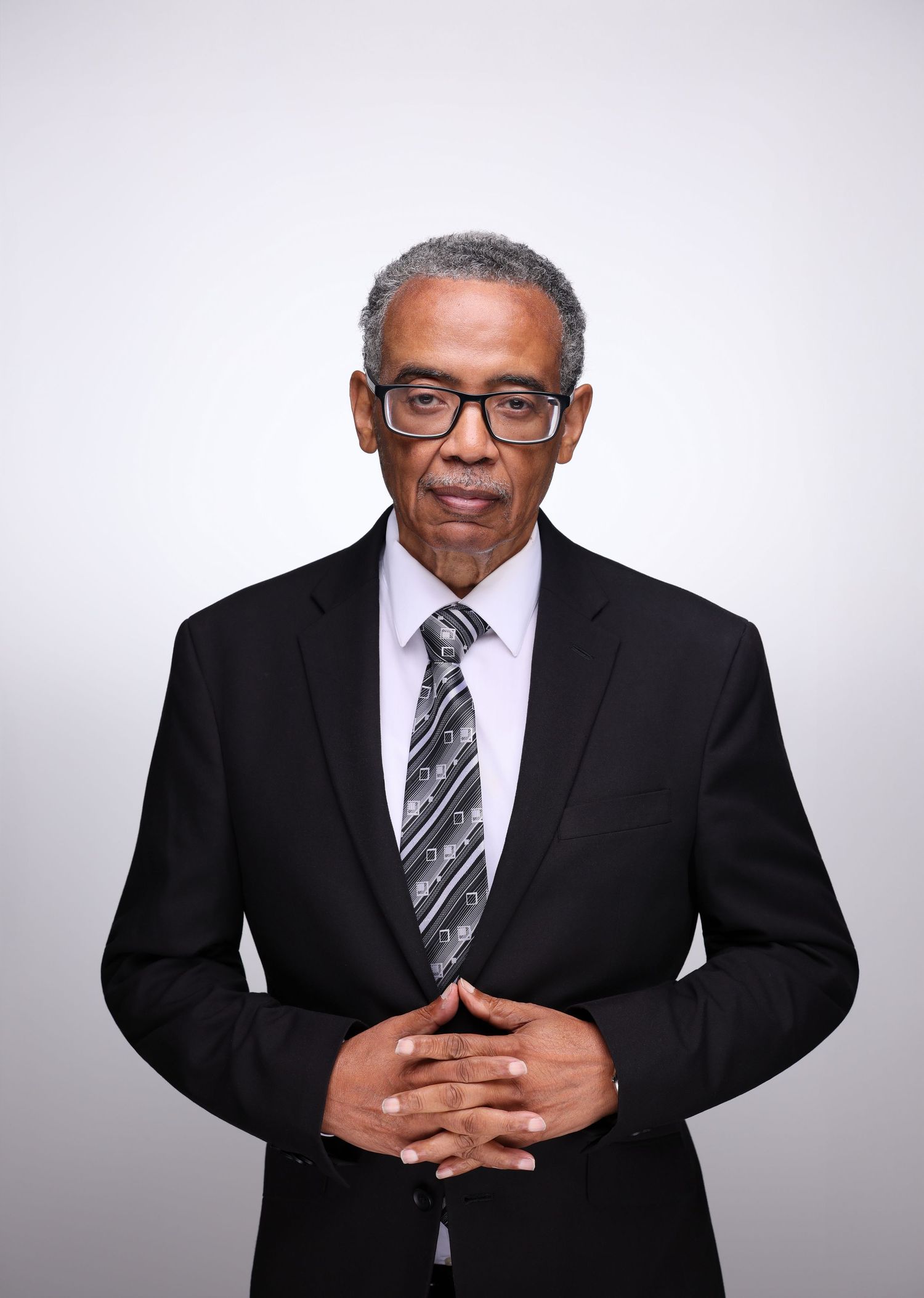 Professional man in black suit and patterned tie, wearing glasses, posing with hands clasped against gray background.