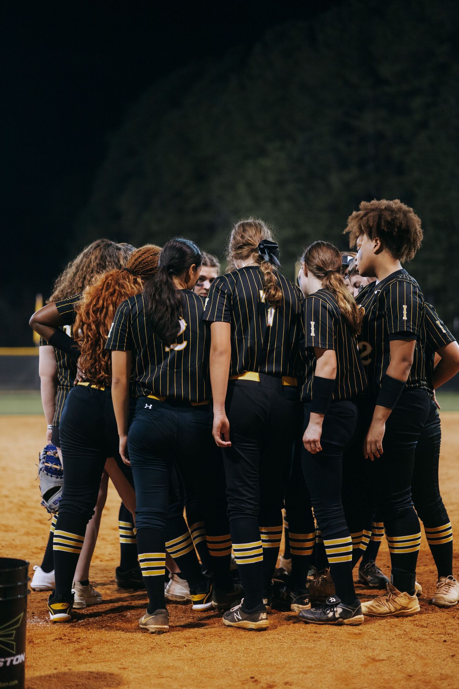 Softball team huddles together on field wearing black and gold pinstriped uniforms.