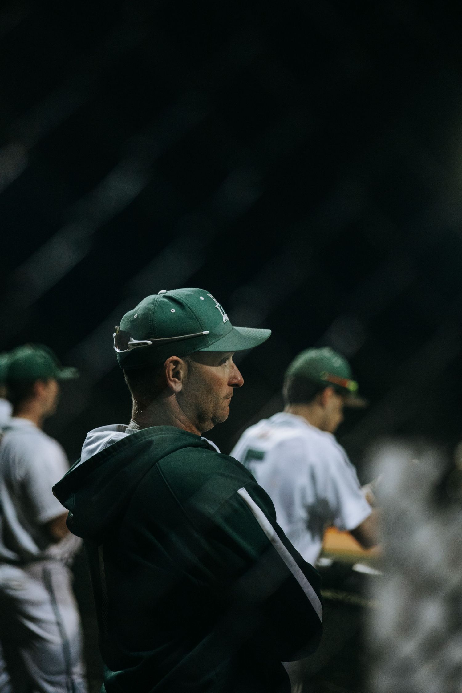 Baseball coach in dark green cap watches from dugout during night game.