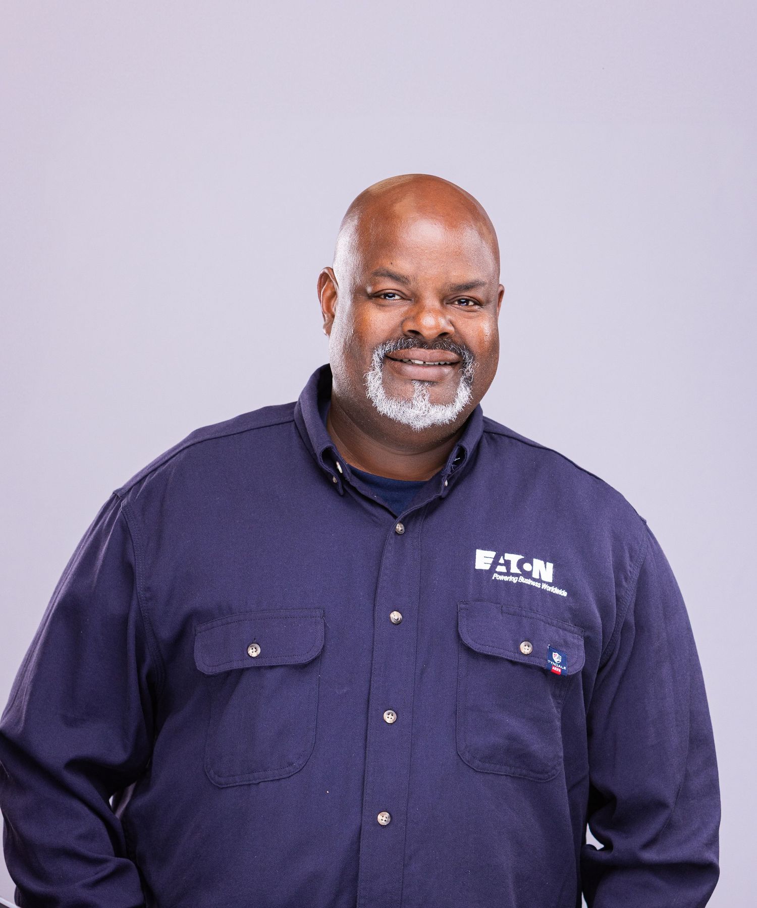 Professional portrait of person in navy work uniform with chest pockets against purple background.