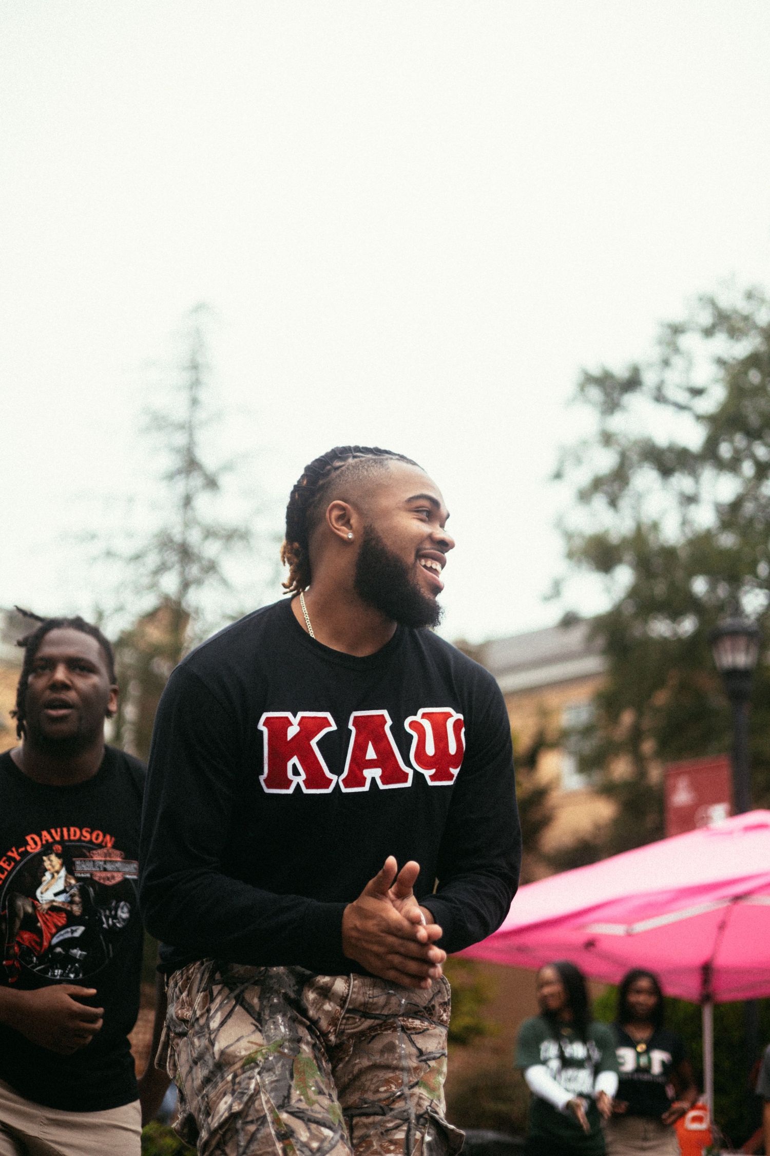 Person wearing a black Kappaa sweatshirt and camouflage pants laughs joyfully outdoors with a pink umbrella visible in background.