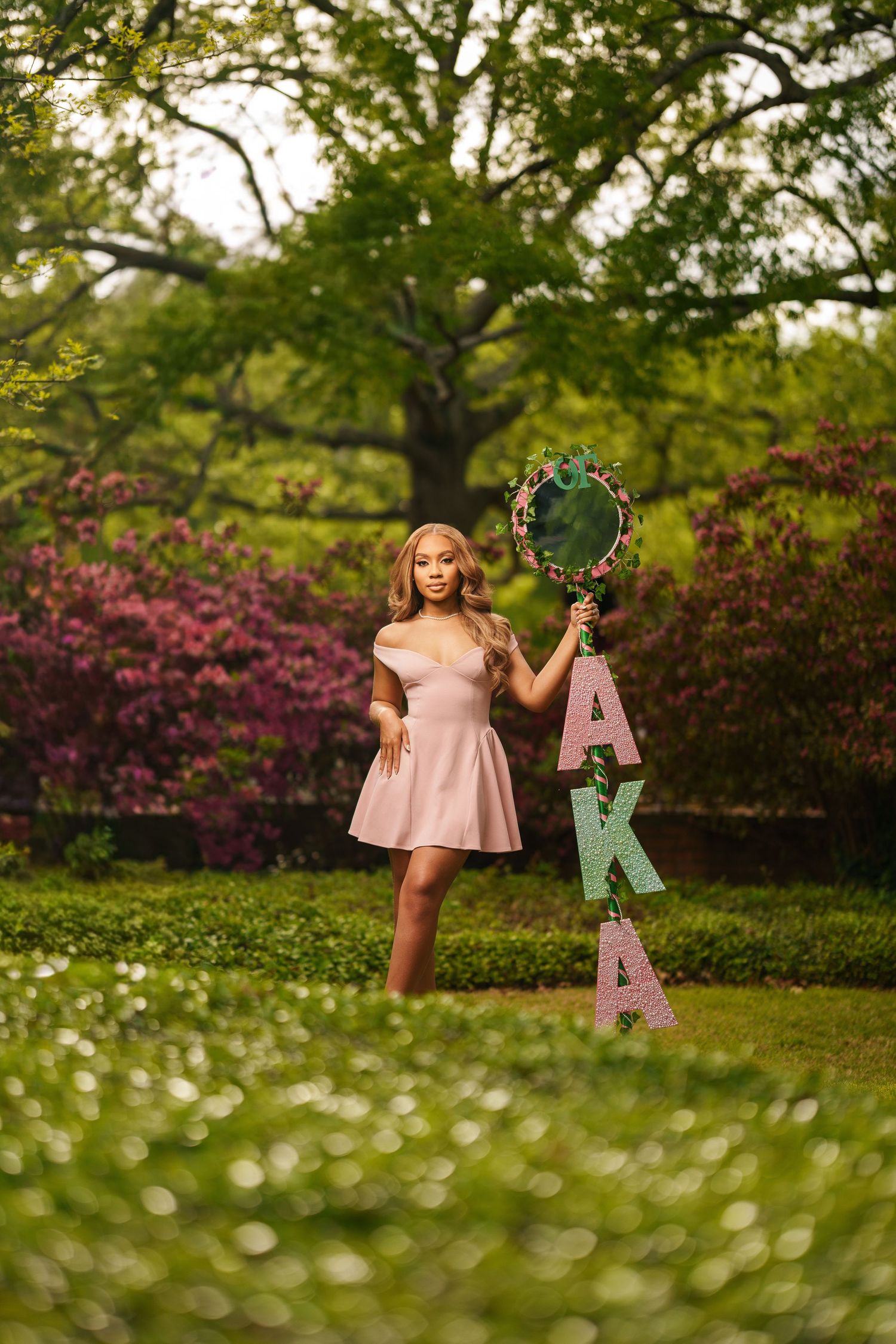 Young woman in pink dress holding AKA sorority letters and mirror outdoors among blooming azaleas and lush greenery.