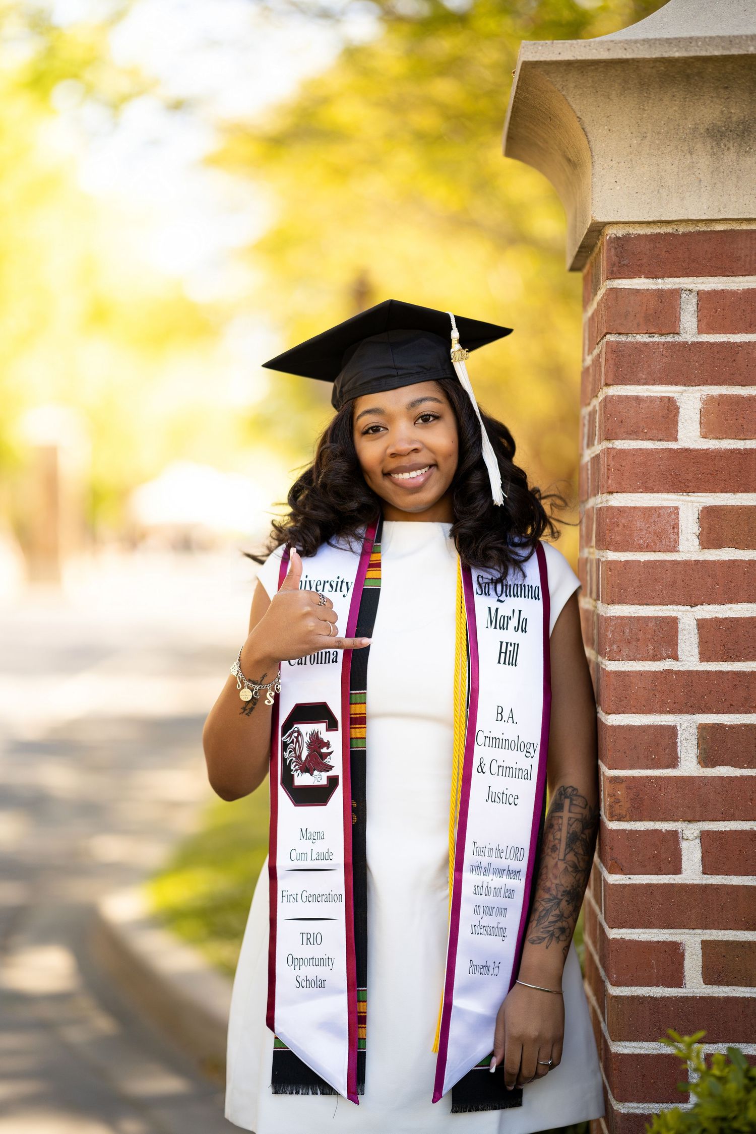 University of South Carolina graduate Mar'Ja Hill smiles in cap, gown, and honor stoles beside a brick column outdoors.