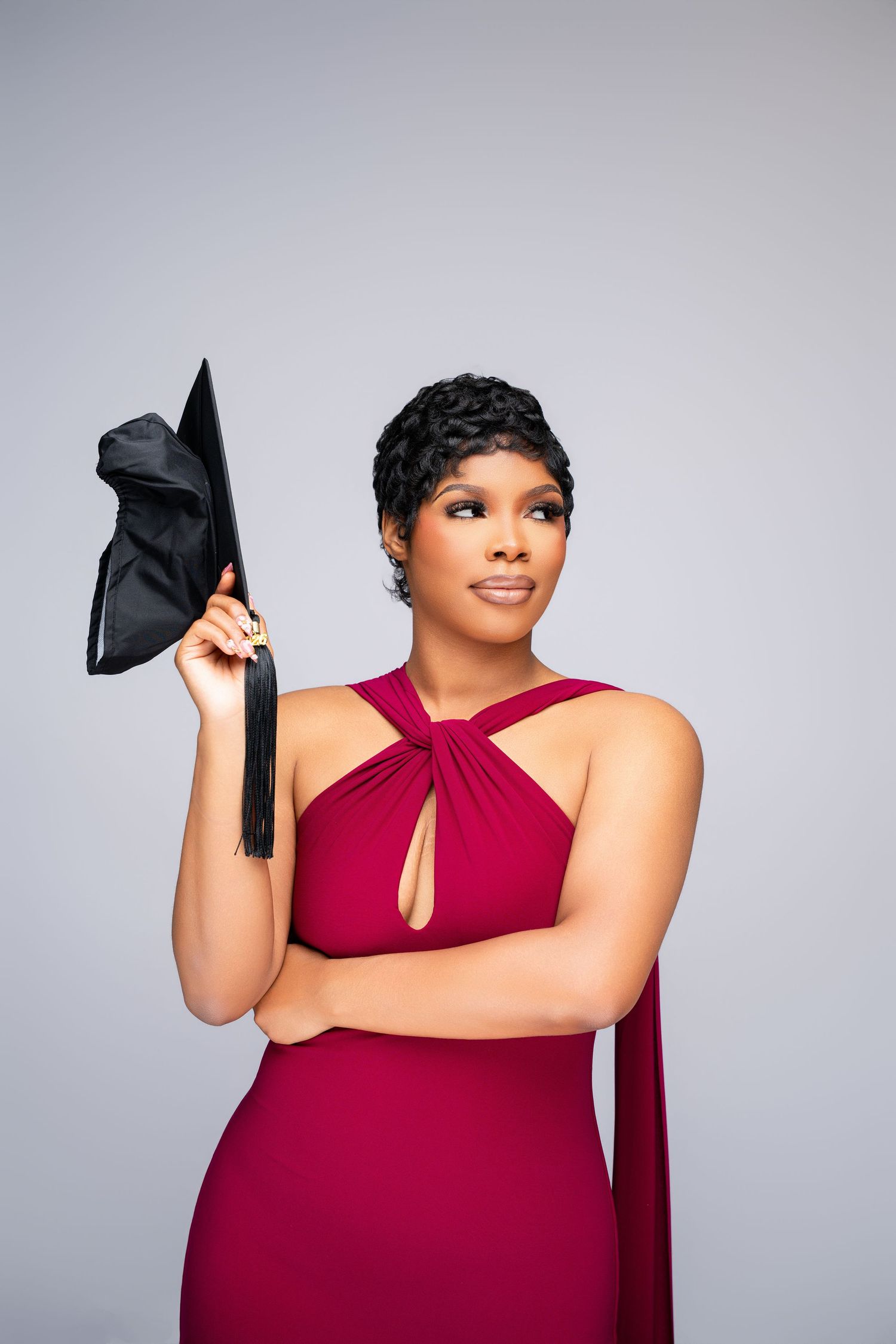 A woman in a burgundy halter gown holds a black graduation cap, posing confidently against a gray background.