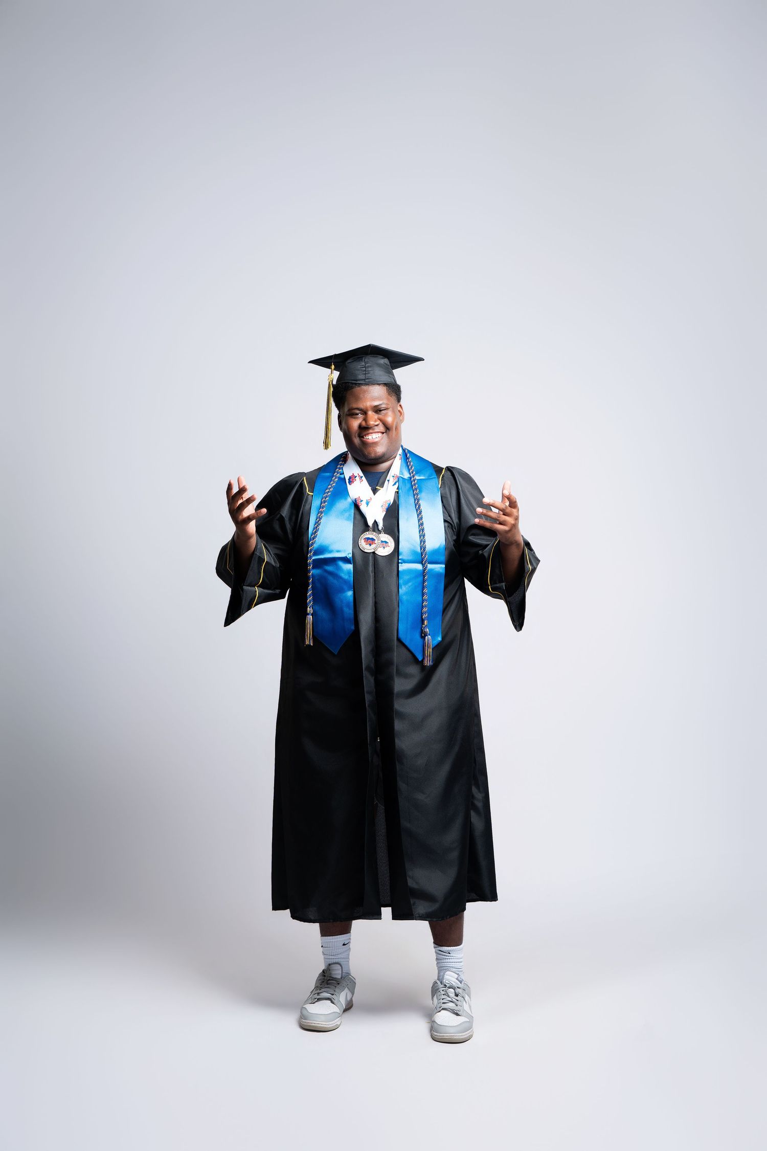 Proud graduate in black cap and gown with blue stole and medal, smiling with hands raised on white background.