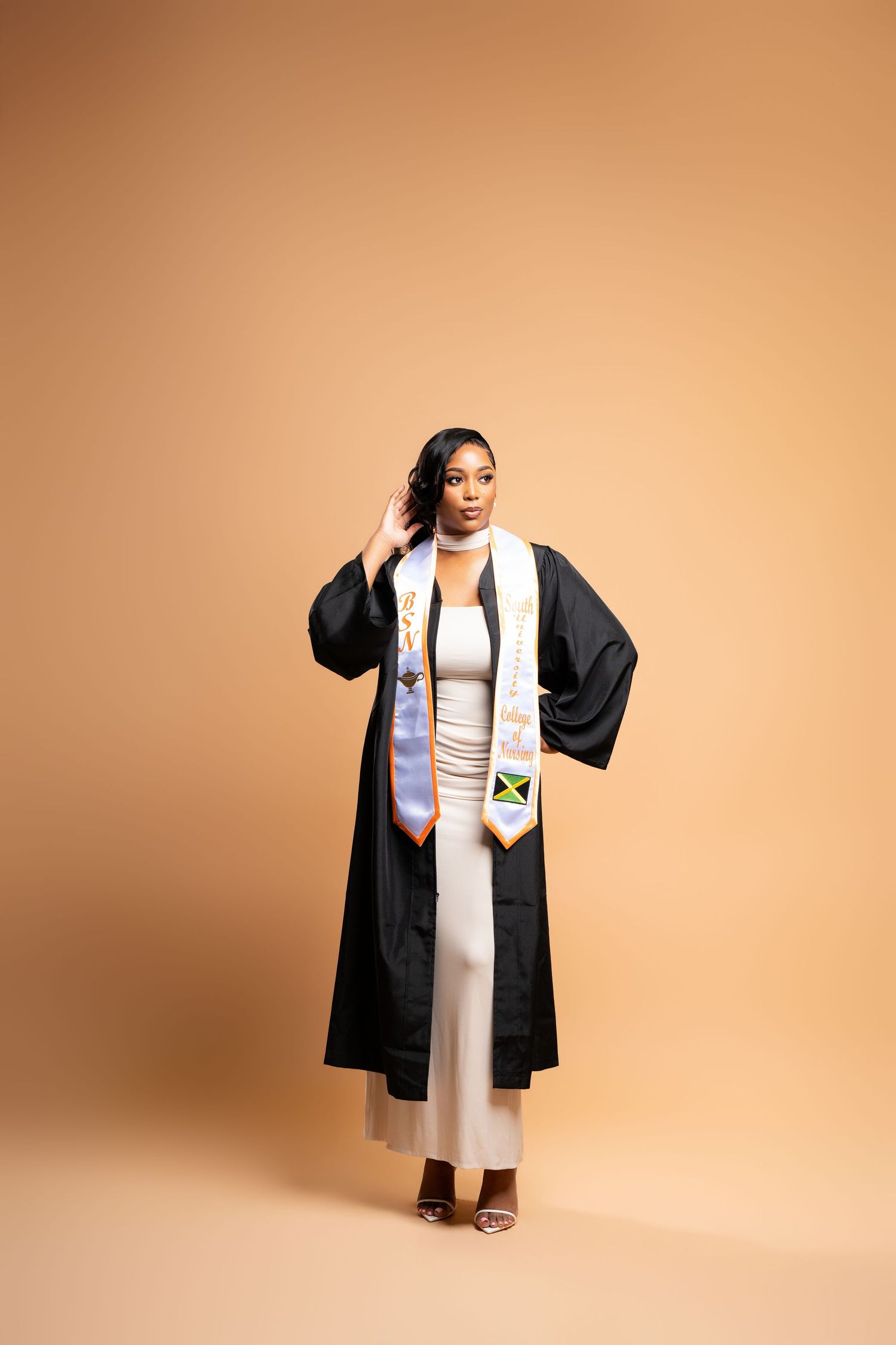 Young woman in black graduation gown and Jamaican flag stole posing confidently against a beige background.
