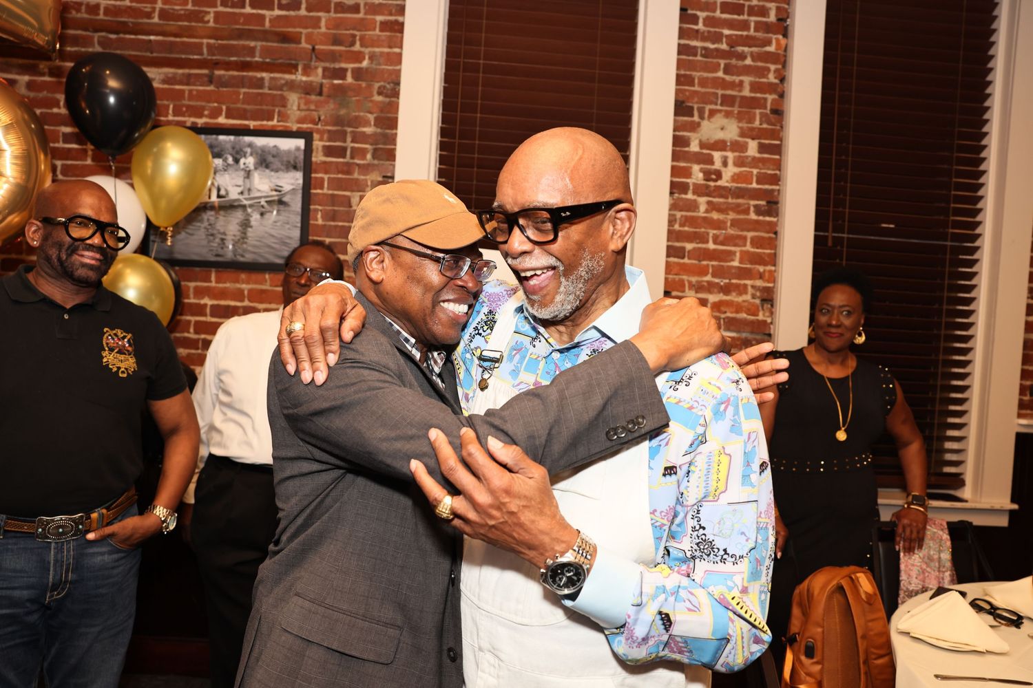 Two men share a warm embrace at a celebratory party with gold and black balloons in the background.
