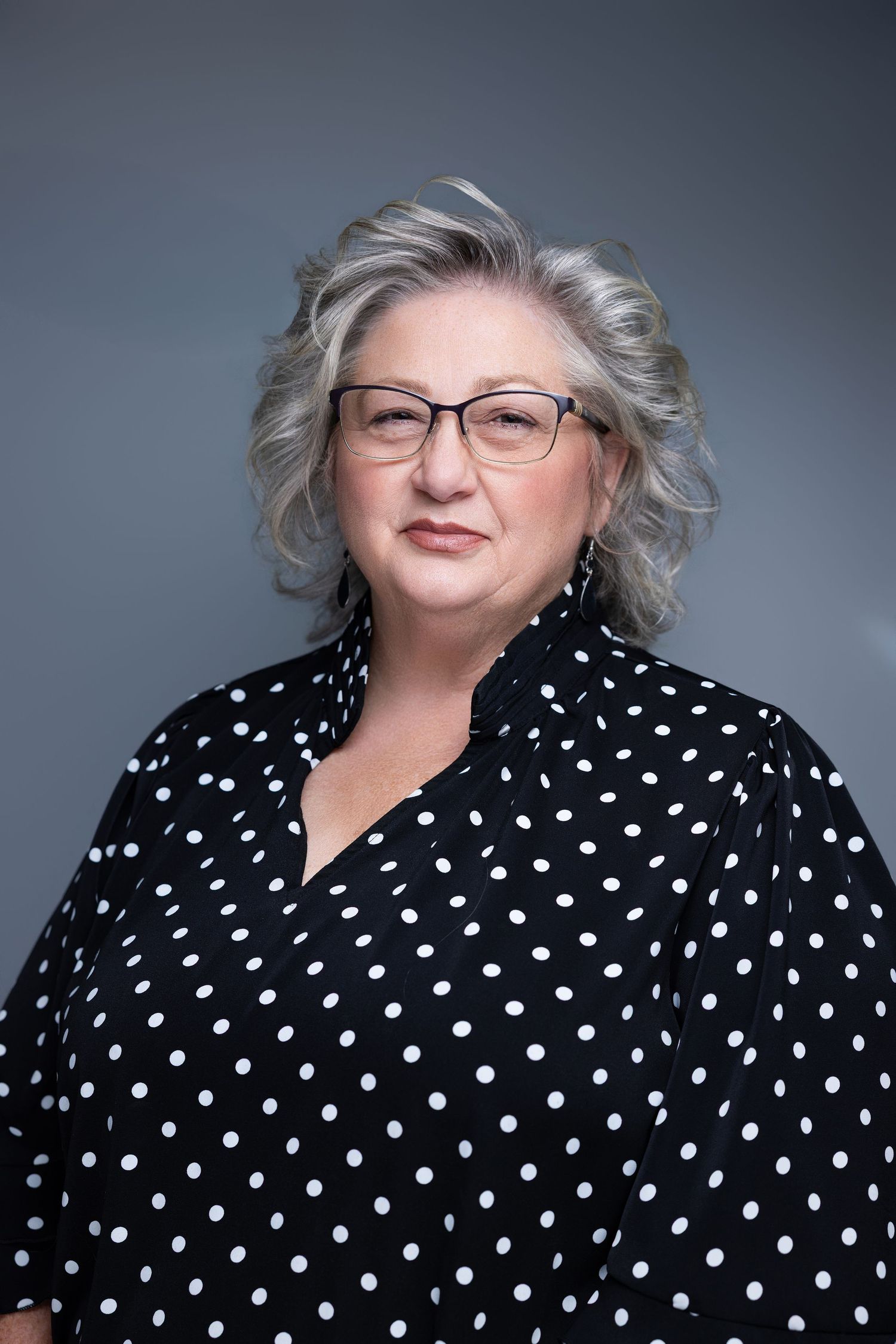 Middle-aged woman with gray hair and glasses wearing a black and white polka dot blouse, professional portrait.