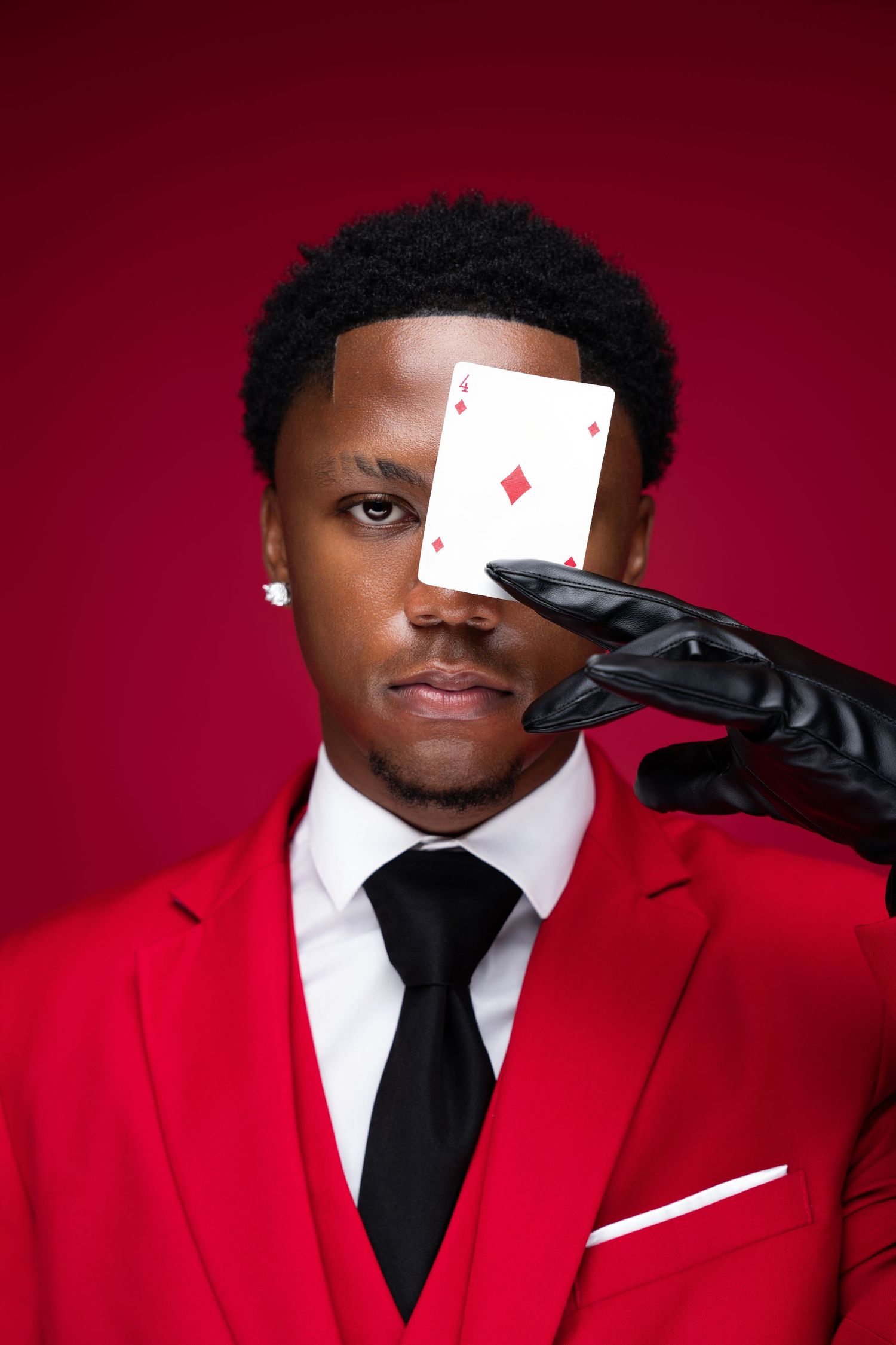 A man in a red suit holds an ace of diamonds playing card over one eye against a red background.