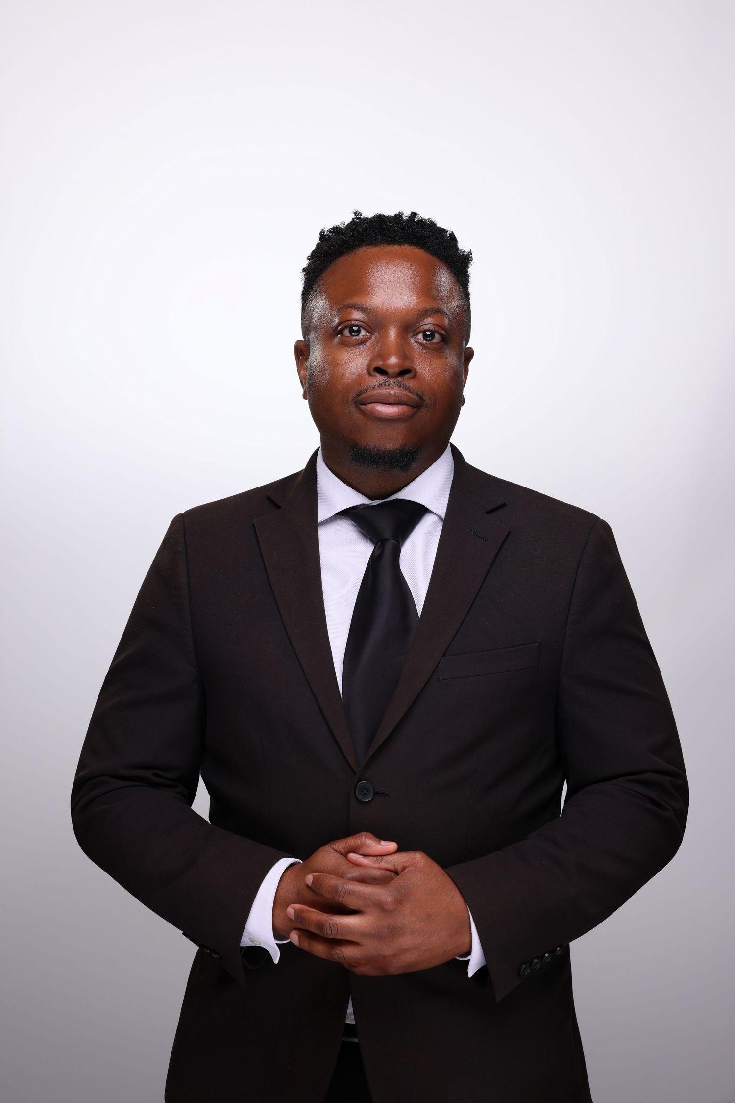 Professional Black man in dark suit and tie with clasped hands posing for formal portrait on gray background.