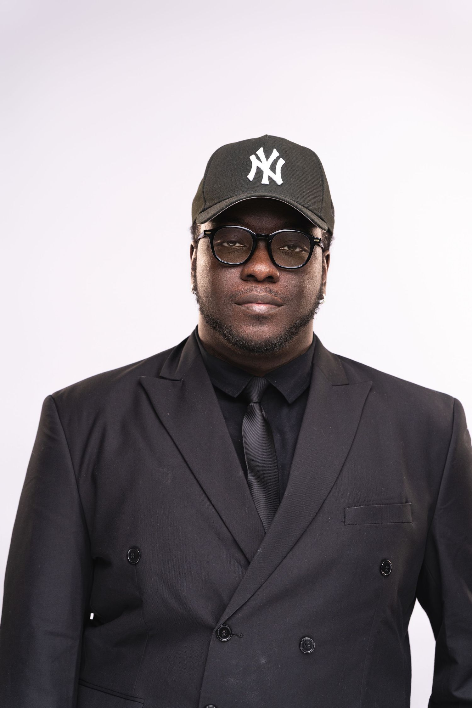 Man in black suit, NY Yankees cap, and glasses posing against white background.